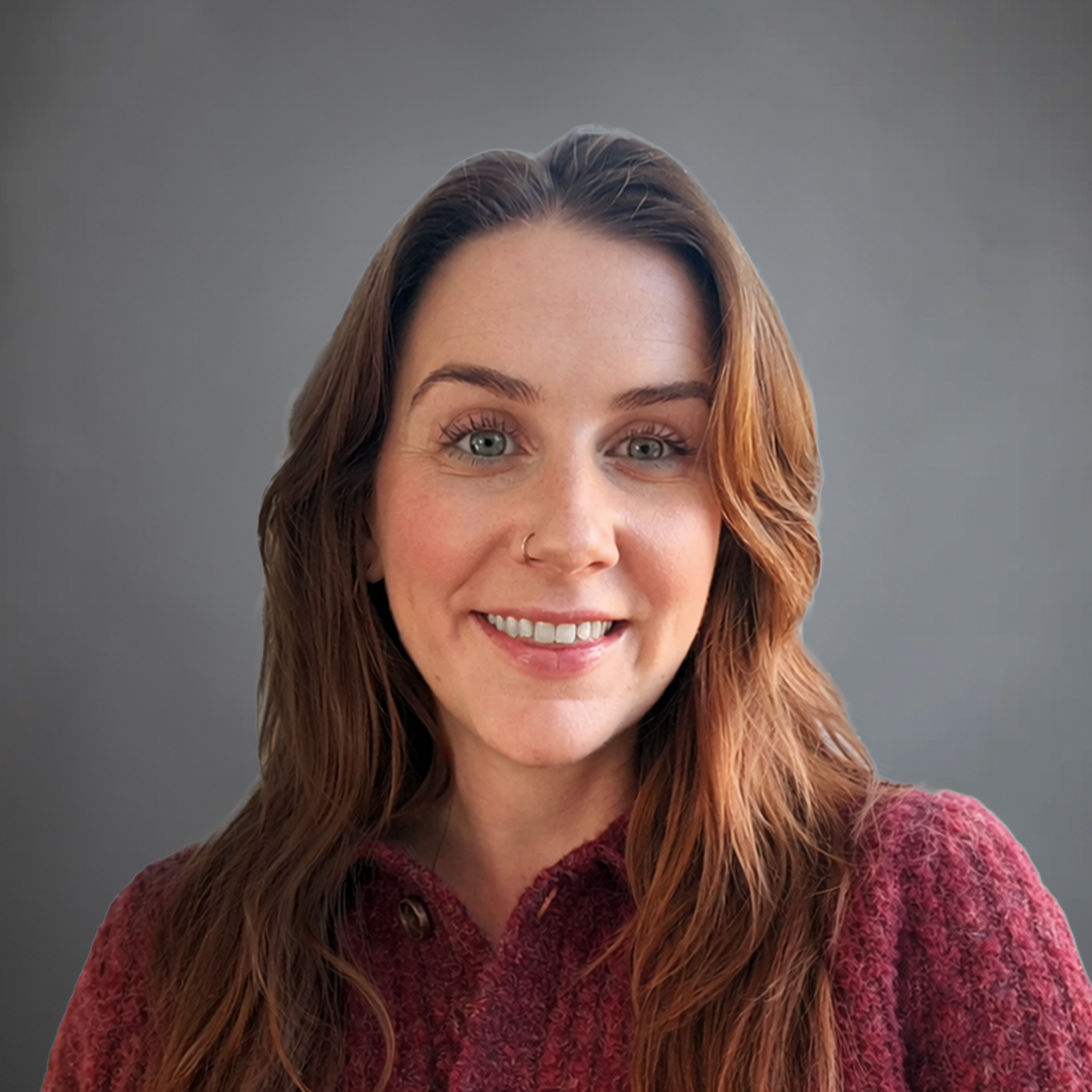 A woman with long wavy brown hair and blue eyes smiles at the camera. She has a nose piercing and is wearing a maroon sweater against a plain gray background.