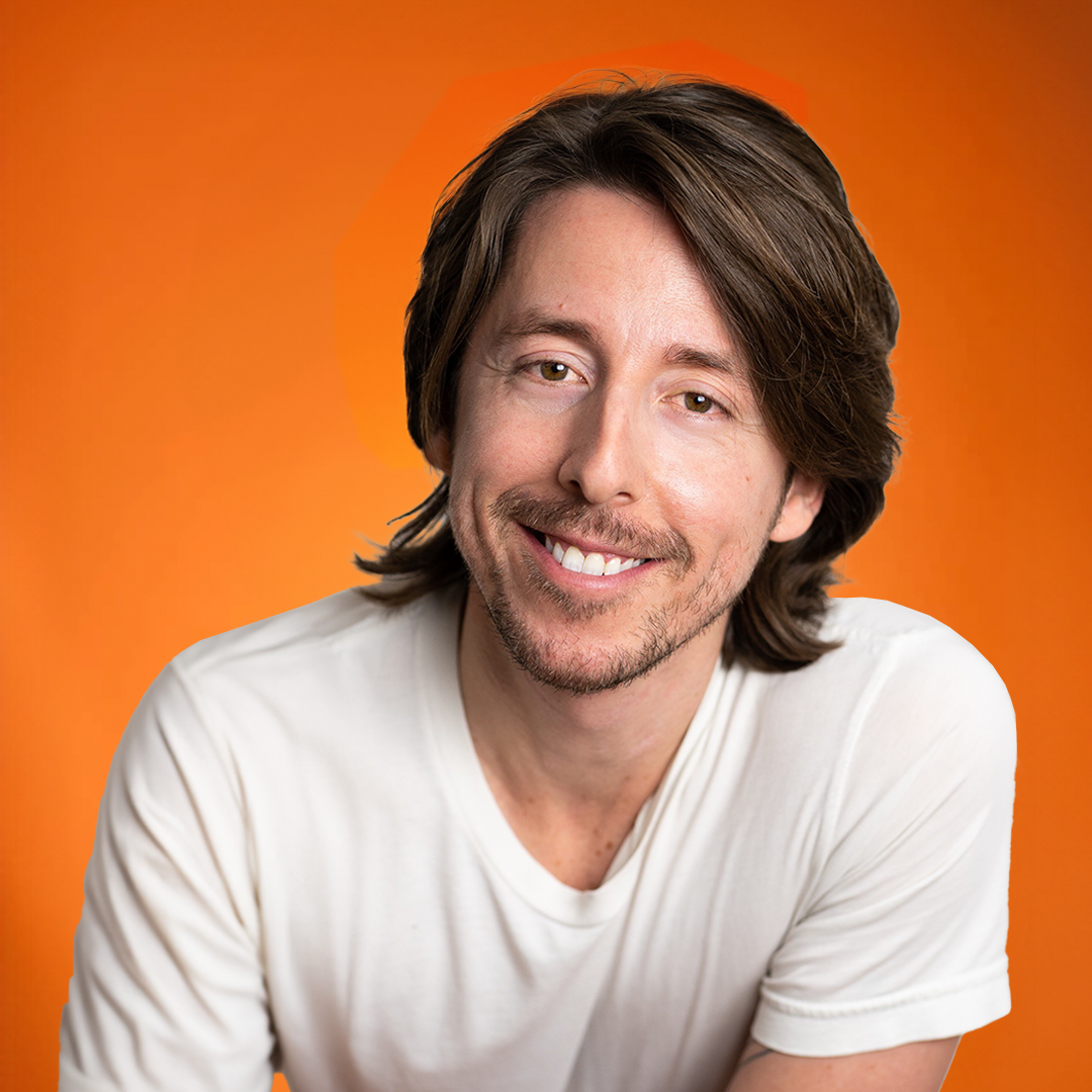 Smiling man with shoulder-length brown hair, mustache, and beard wearing a white t-shirt against an orange background.