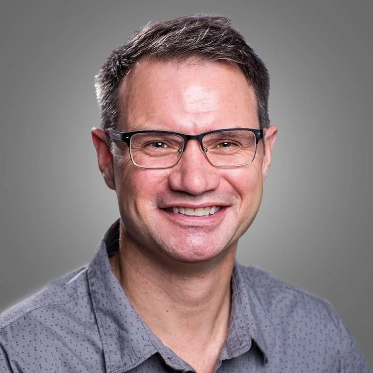 A smiling man wearing glasses and a gray, patterned shirt.