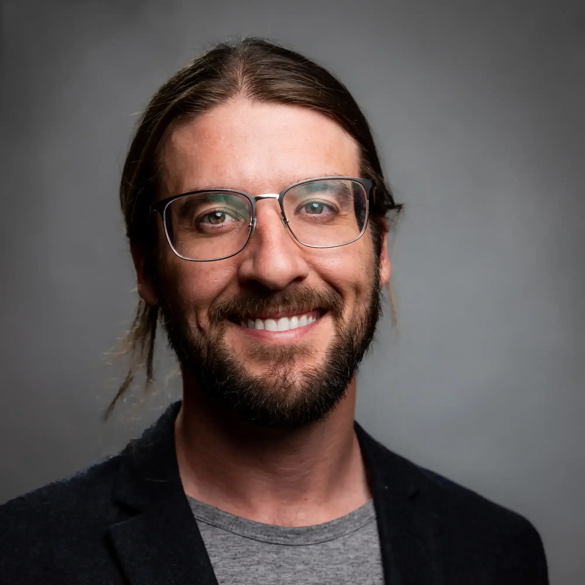 A smiling man with glasses, a beard, and long brown hair tied back, wearing a black jacket and a gray shirt, against a neutral gray background.