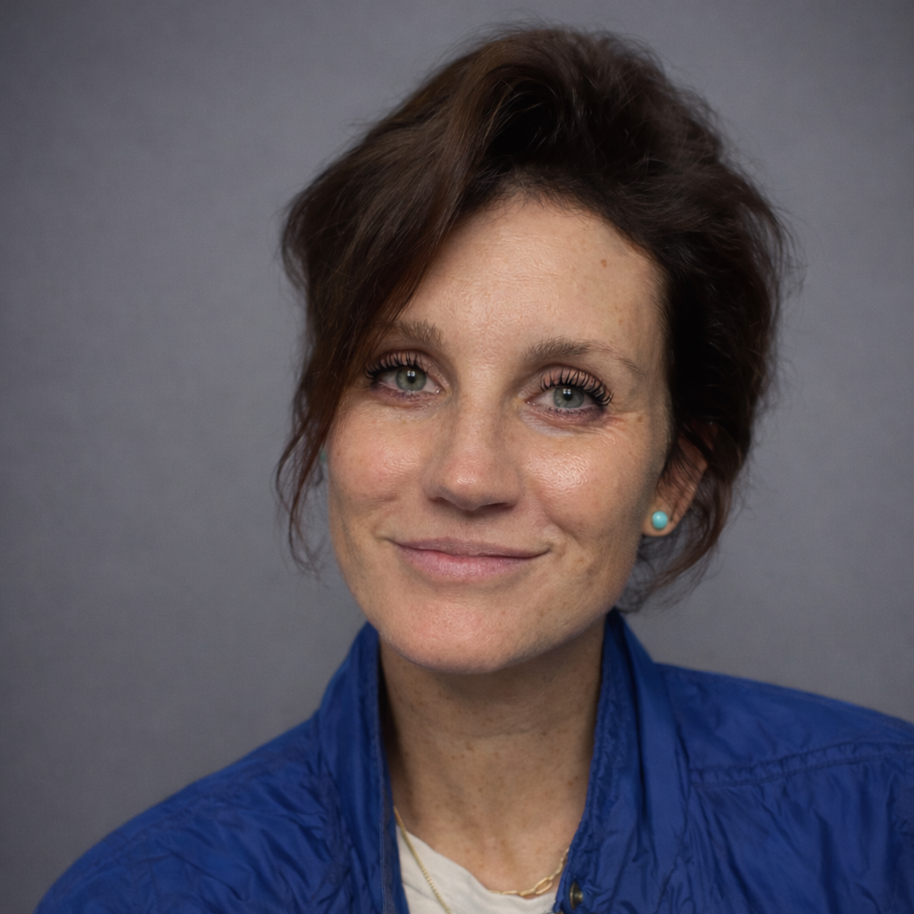 Close-up of a woman with short, wavy brown hair and green eyes, wearing a blue jacket and small turquoise earrings, smiling at the camera against a gray background.