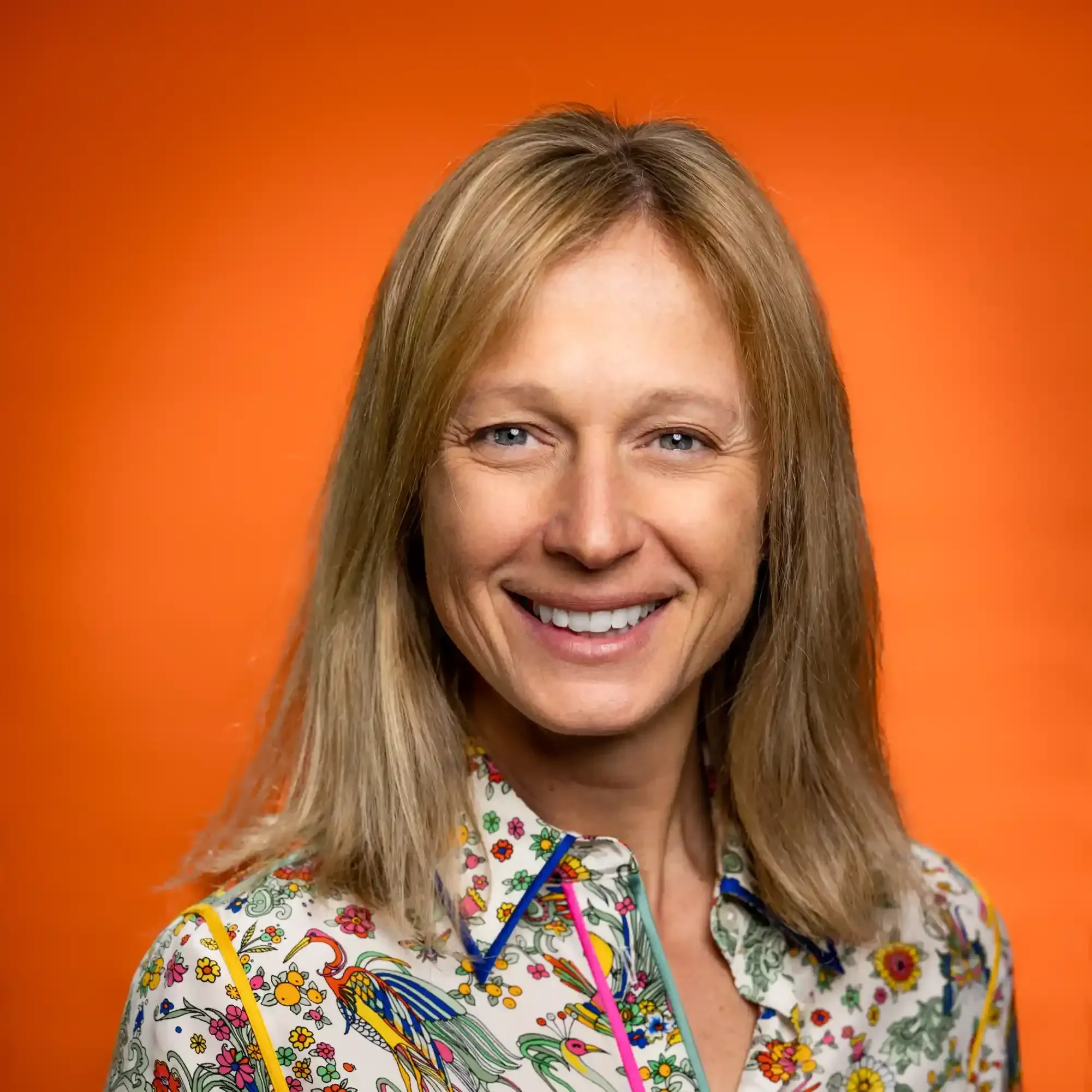 A smiling woman with shoulder-length blond hair and blue eyes, wearing a colorful, floral-patterned shirt against an orange background.
