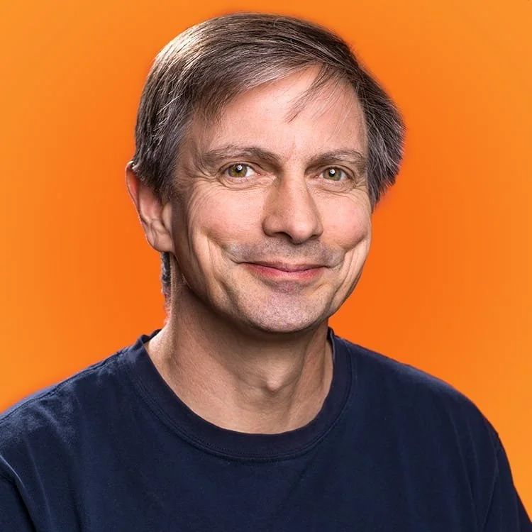 Portrait of a smiling man with gray hair, wearing a dark blue shirt, against an orange background.