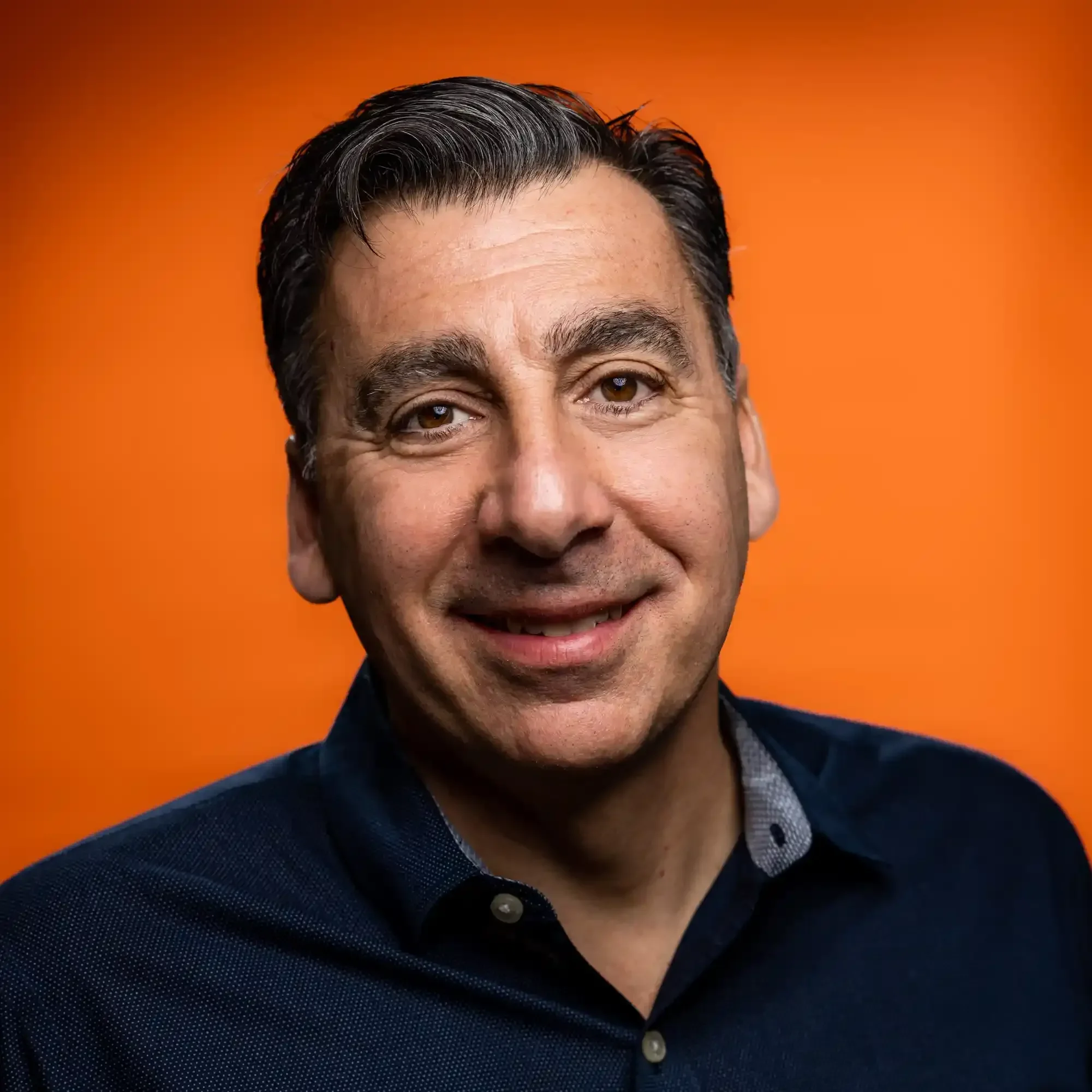 Headshot of a smiling man with dark hair and brown eyes, wearing a dark shirt against an orange background.