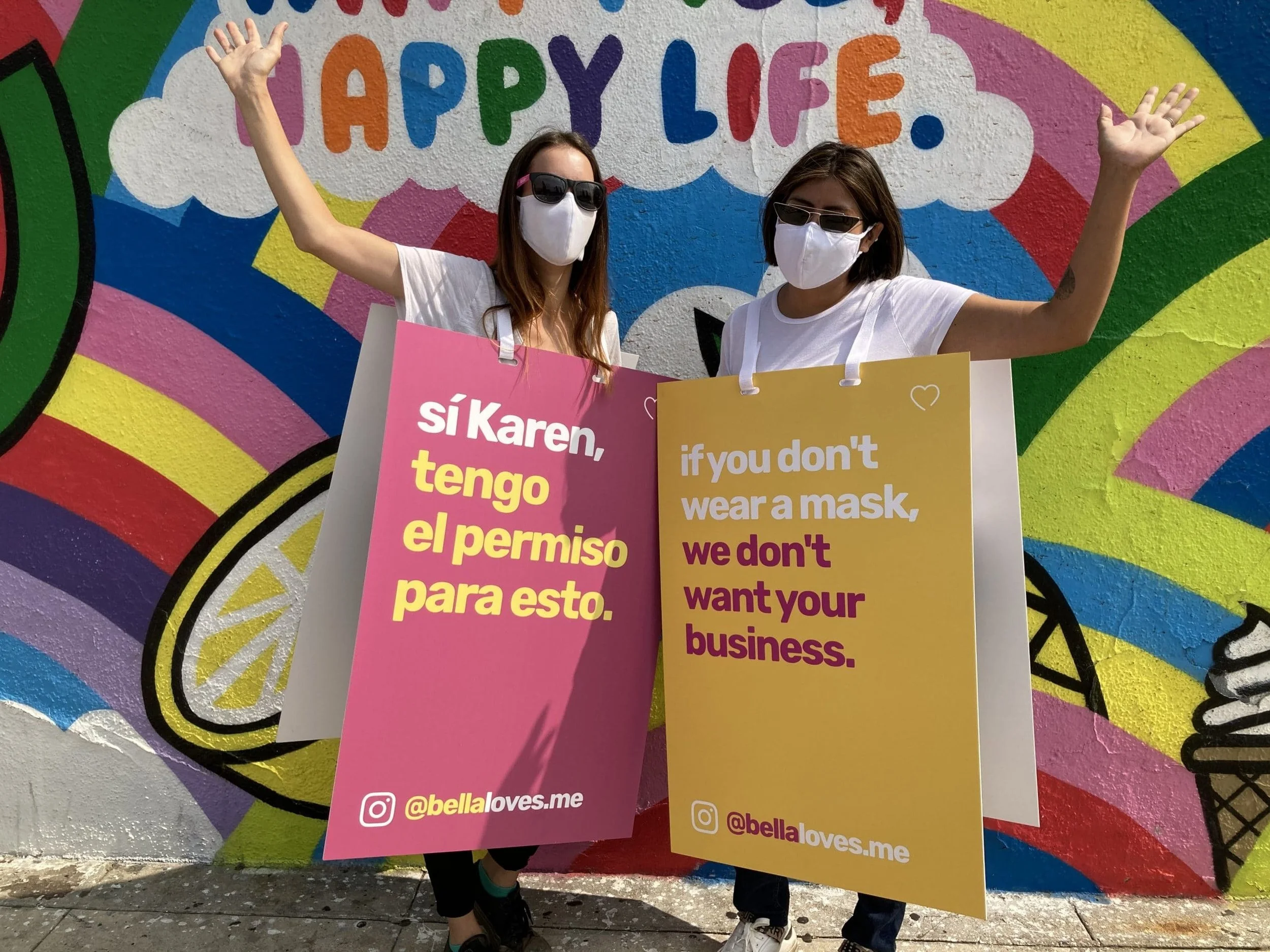 Two women wearing face masks and white sandwich boards with messaging about wearing masks stand waving in front of a colorful rainbow mural reading "Happy Life."