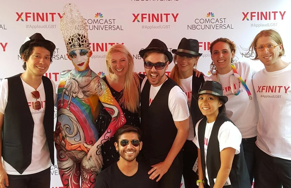 A group posing in front of an Xfinity #ApplaudLGBT backdrop alongside a performer in full-body paint and an ornate headpiece.