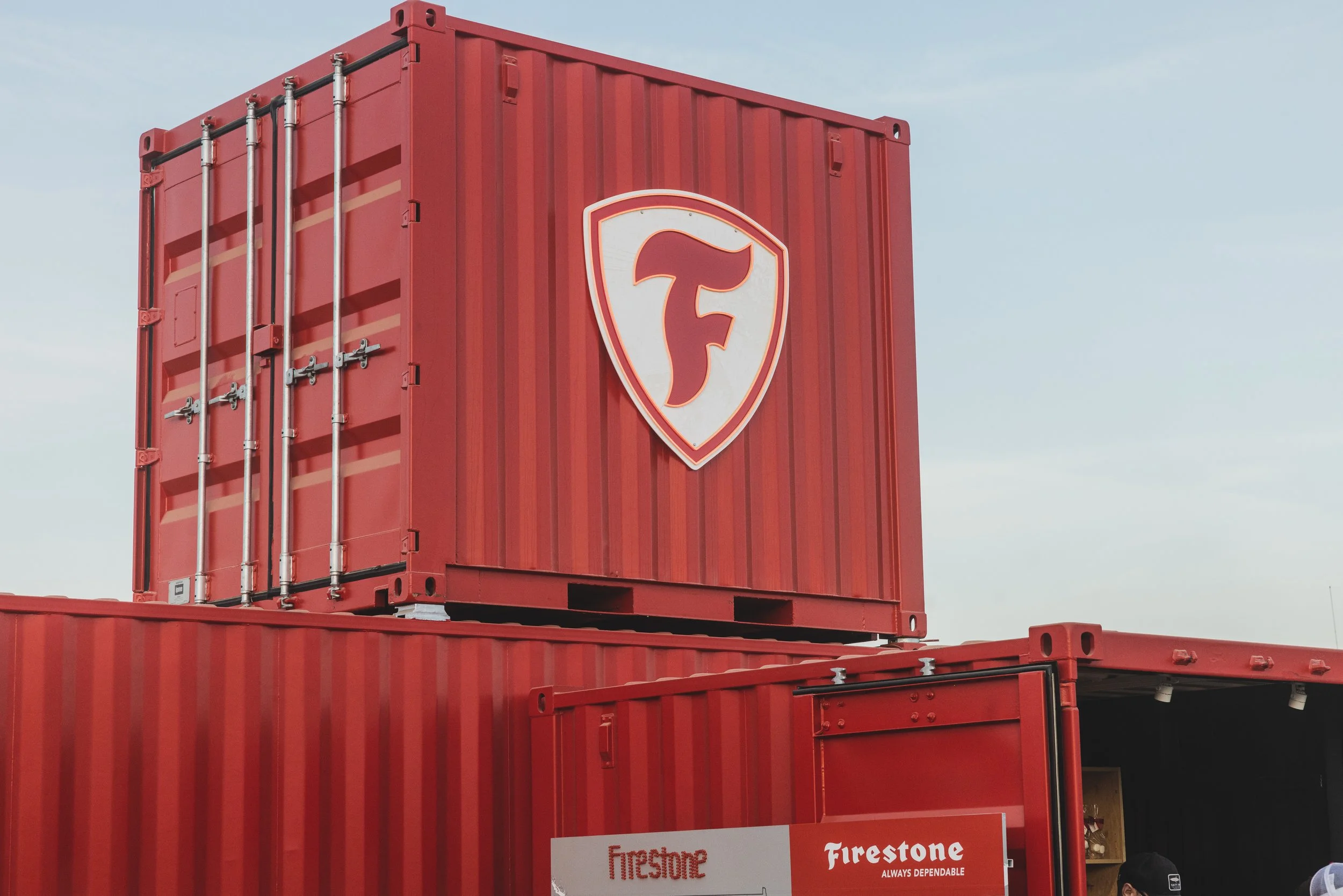 A red shipping container with the Firestone logo on it, stacked among other similar containers. The sky is partly cloudy in the background.
