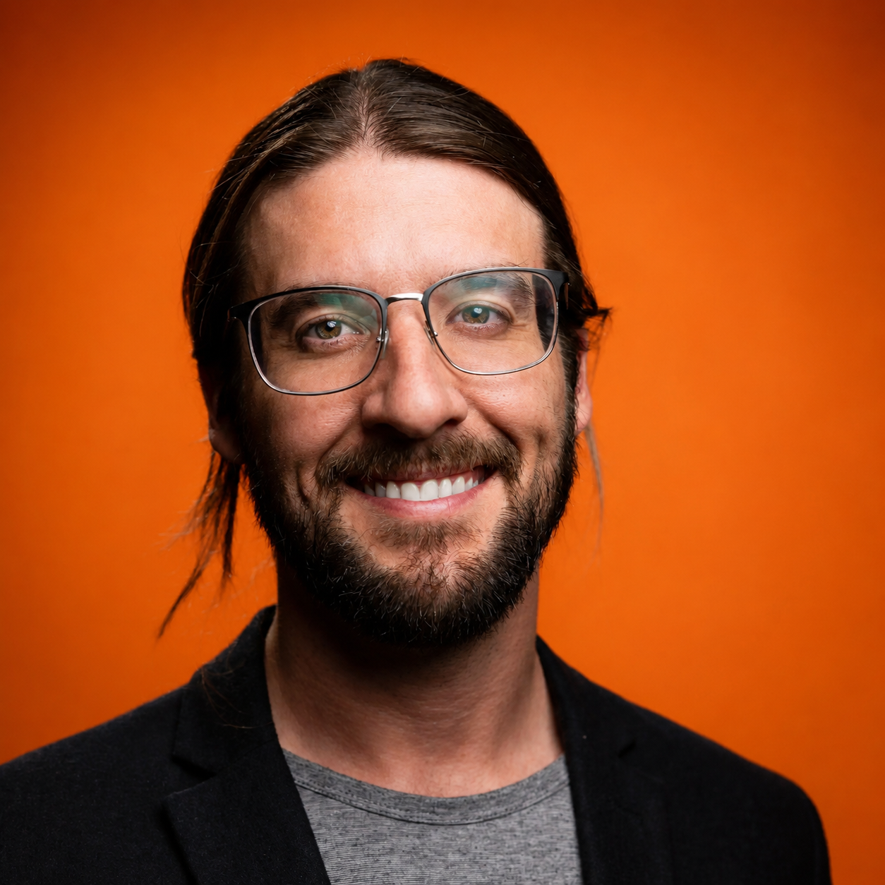 Smiling man with glasses, beard, and long hair against an orange background.