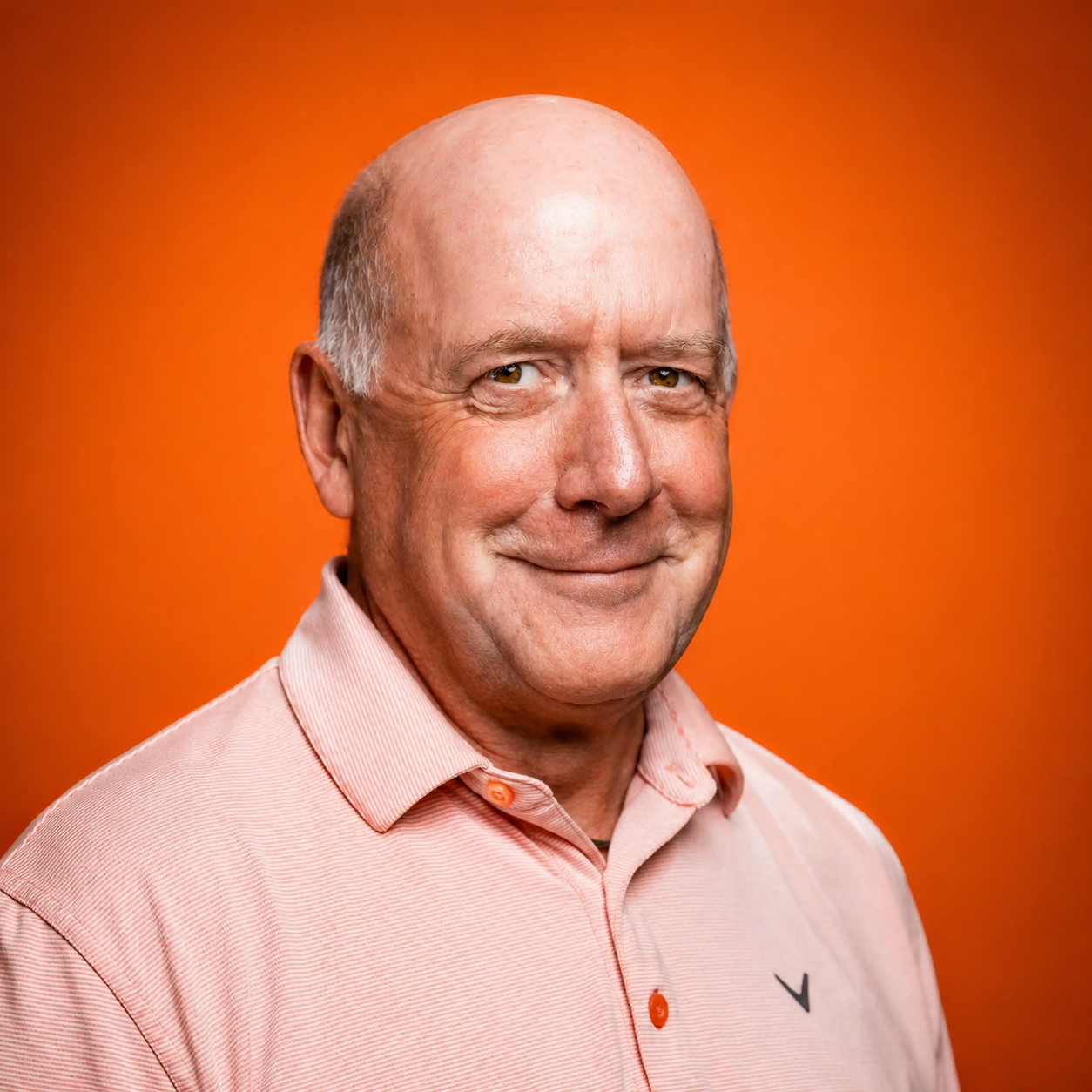 An older man with a bald head, smiling, wearing a light pink button-up shirt, against a solid orange background.