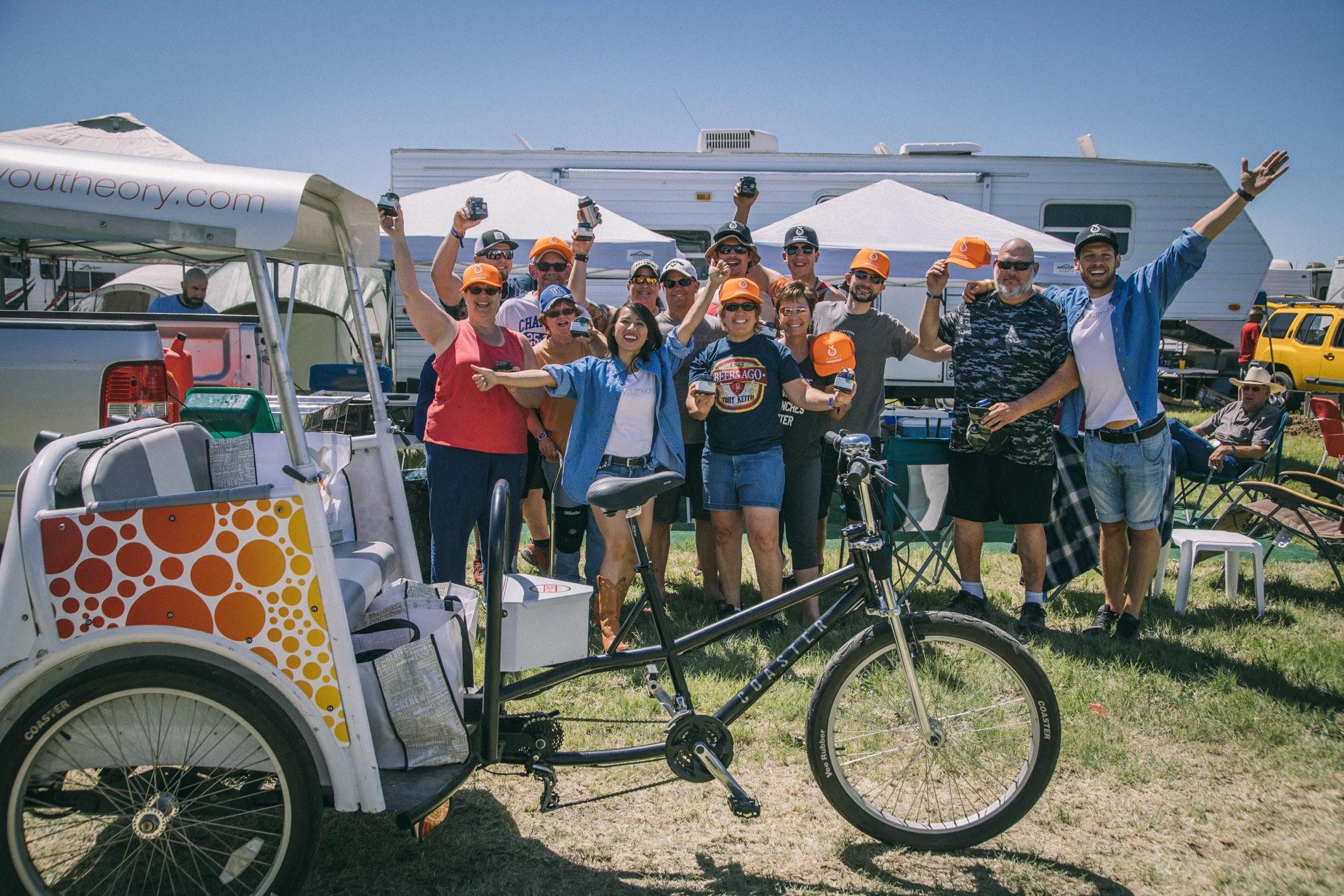 A group of people smiling and celebrating outdoors at a gathering, with some holding small trophies or awards, in a campsite area with RVs and tents, and a bicycle in the foreground.