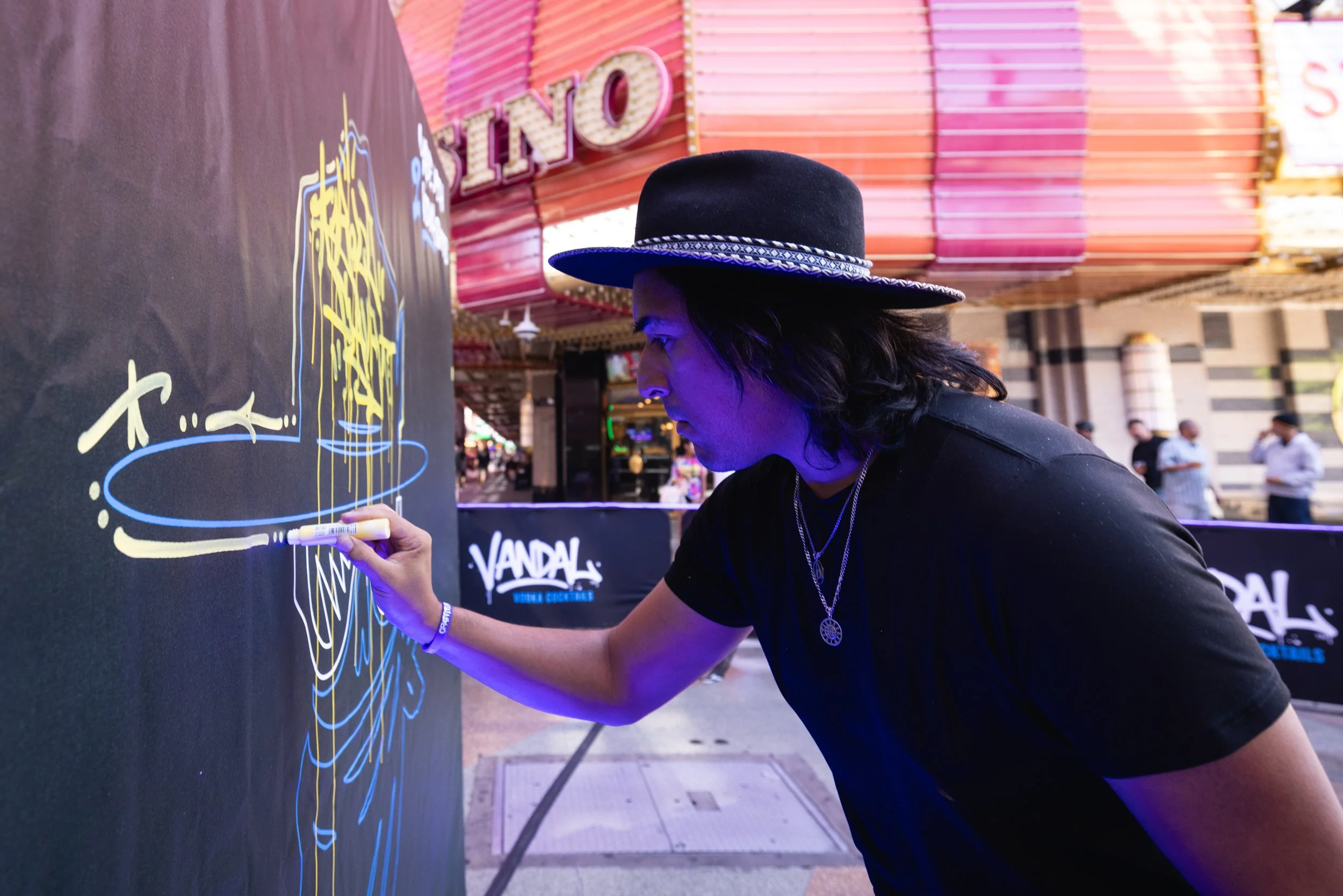 A person wearing a black hat with a decorative band is drawing or writing with a white marker on a large blackboard or poster at an outdoor event. The background features a pink and orange marquee with the word 'NO' visible and a few people walking nearby.