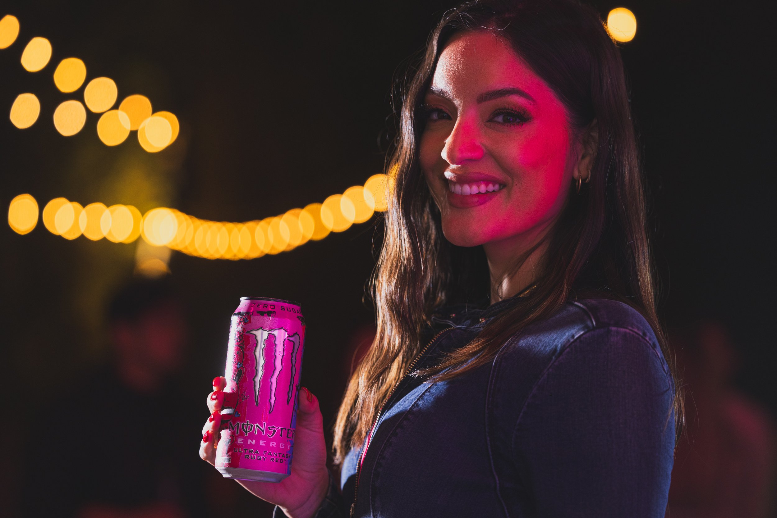 A young woman smiling at night while holding a pink Monster Energy drink, illuminated by warm bokeh lights in the background, wearing a black leather jacket with long wavy hair.