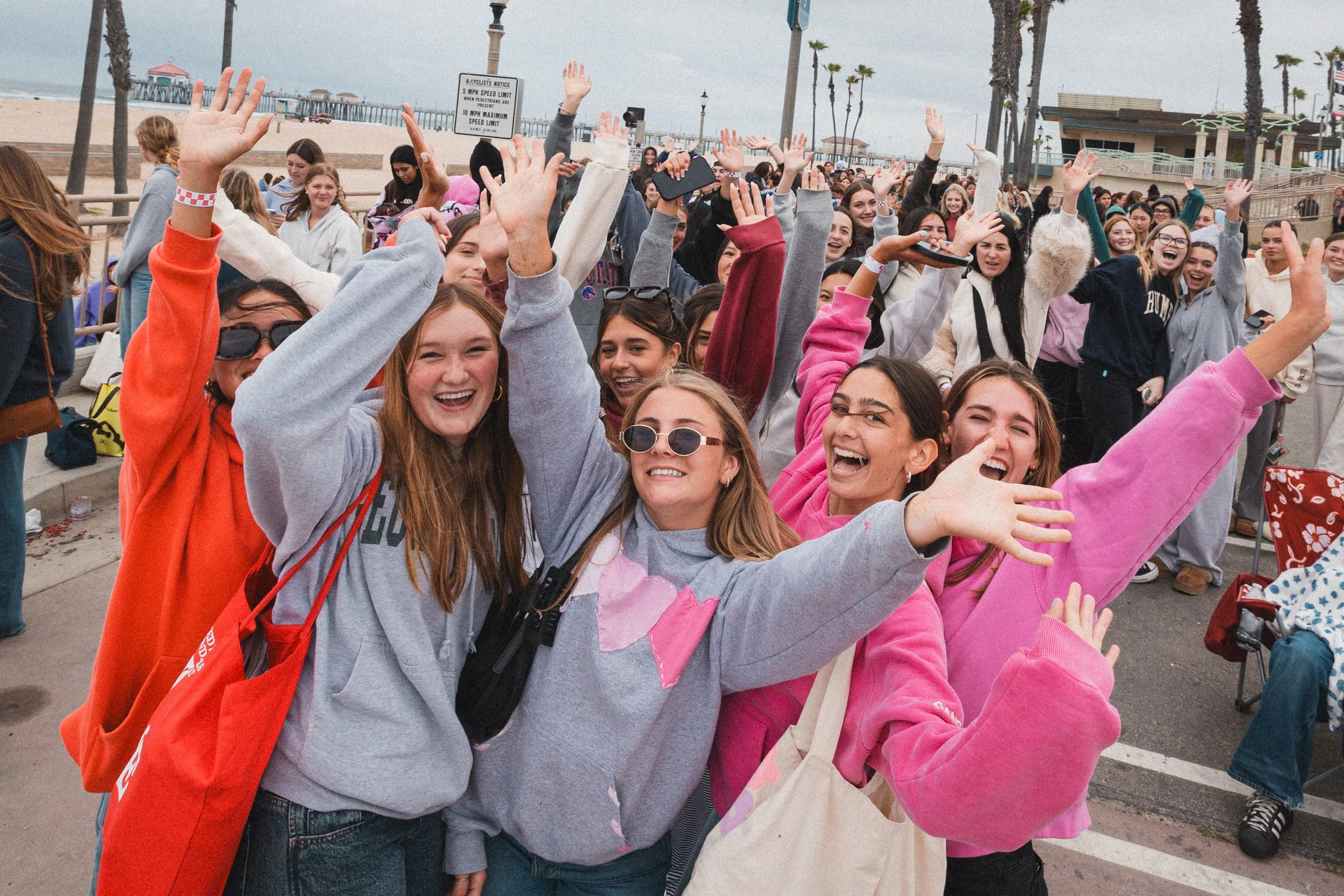 Group of young women and girls at the beach, smiling, raising hands, and enjoying a sunny day near the pier with palm trees in the background.