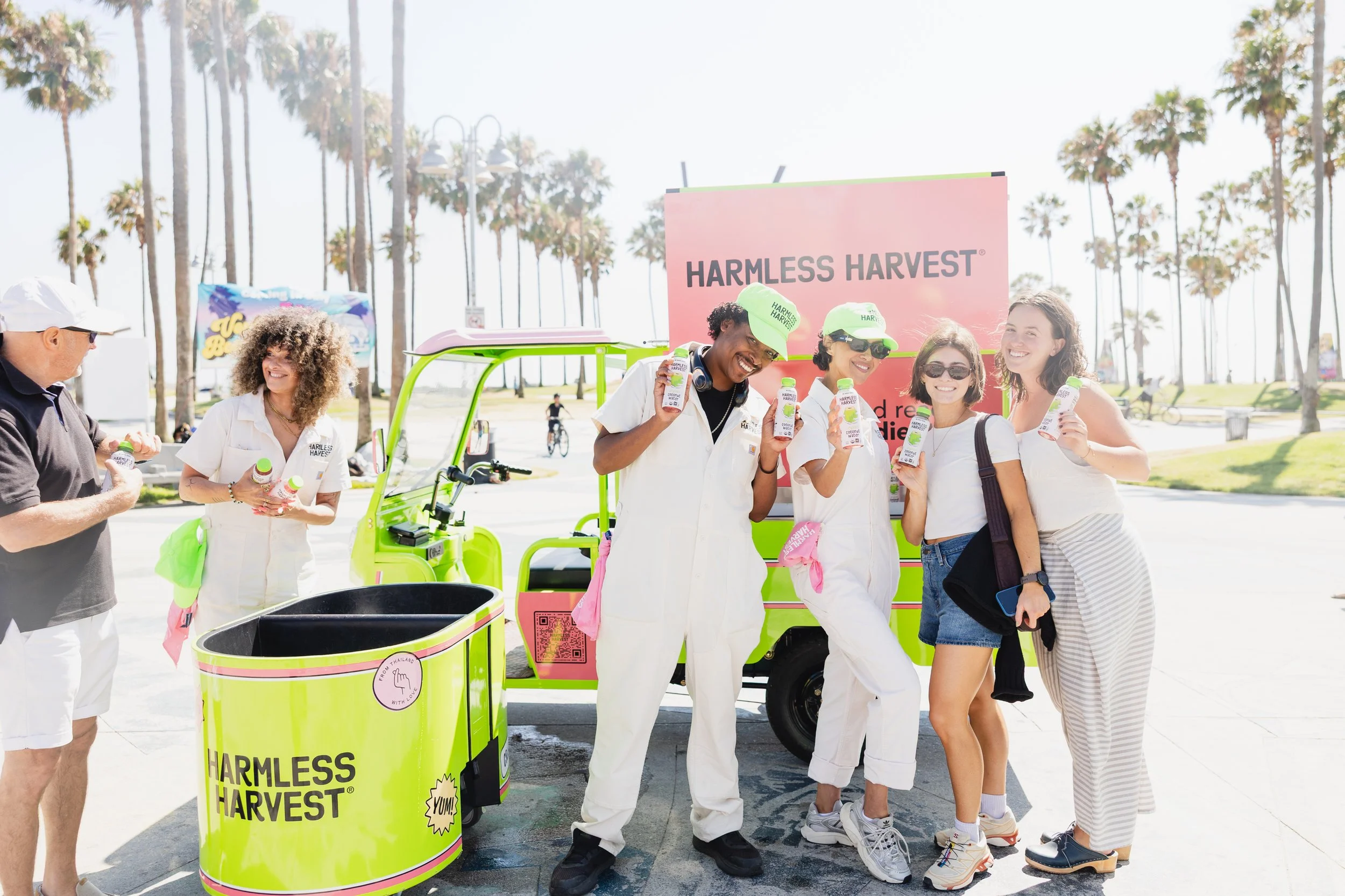 Group of people at a Harmless Harvest smoothie stand on a sunny day with palm trees in the background, holding bottles of coconut water and smiling.