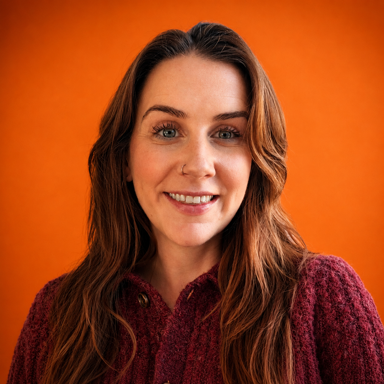 A woman with long wavy brown hair and blue eyes smiling, wearing a maroon textured sweater, against an orange background.