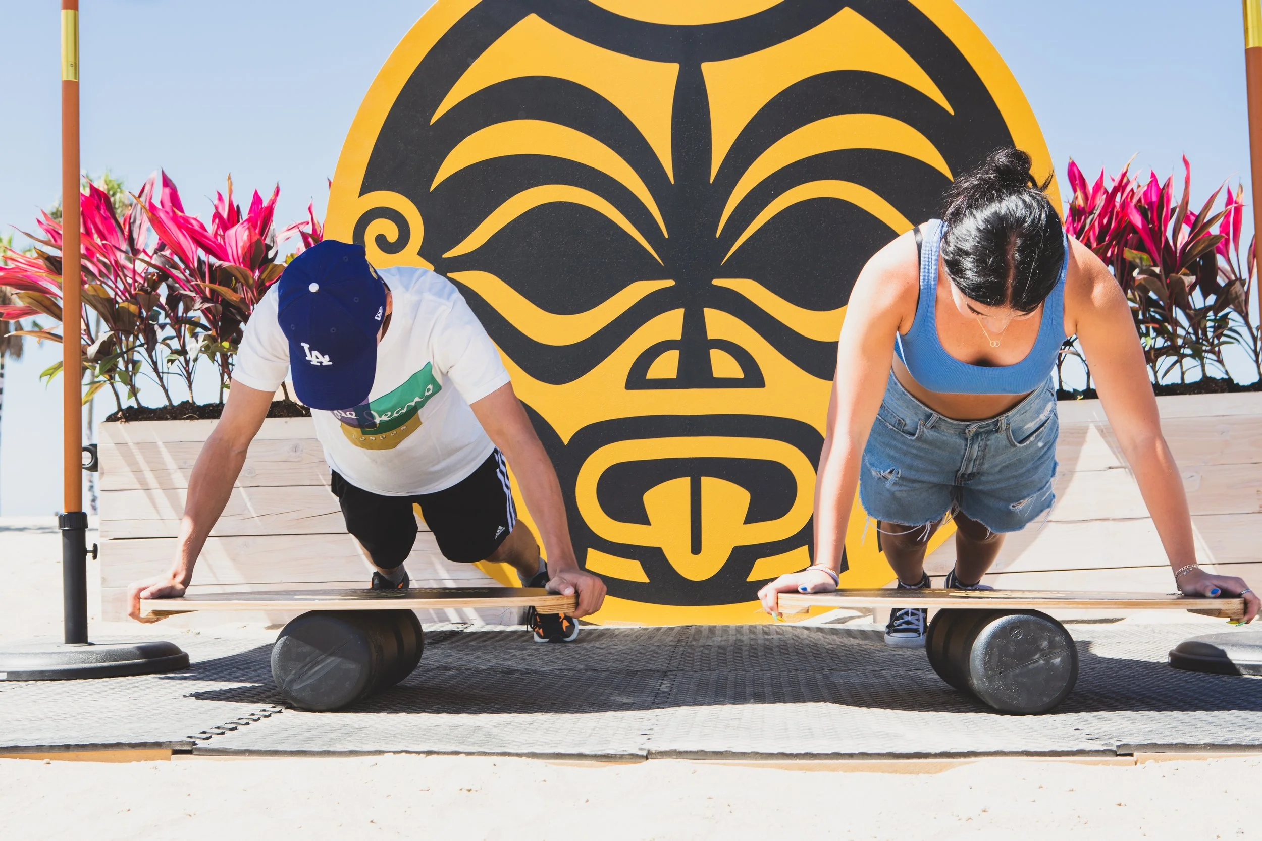 Two people are doing push-ups outdoors in front of a yellow and black tiki-style face sign, surrounded by pink and green plants and a clear blue sky.