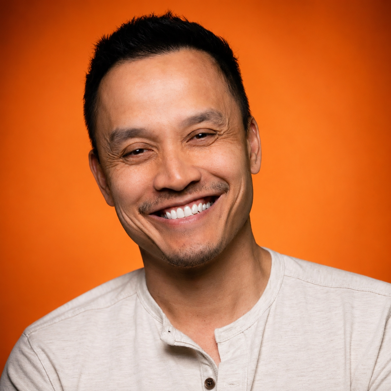 A man smiling with a close-cropped haircut, wearing a light gray shirt, against an orange background.