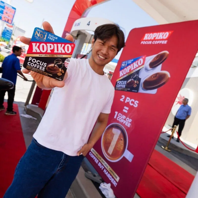 A smiling man holding a Kopiko Focus Coffee candy in front of a promotional sign at an outdoor event, with other people and tents in the background.
