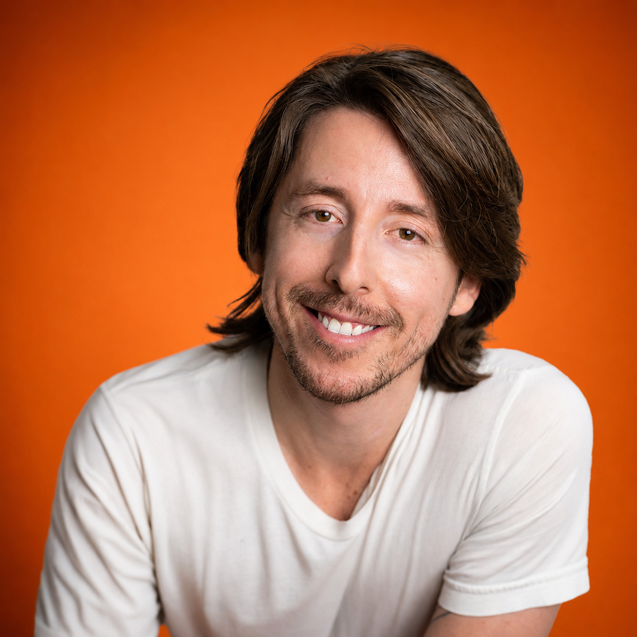 Portrait of a smiling man with brown hair and a beard, wearing a white t-shirt, against an orange background.