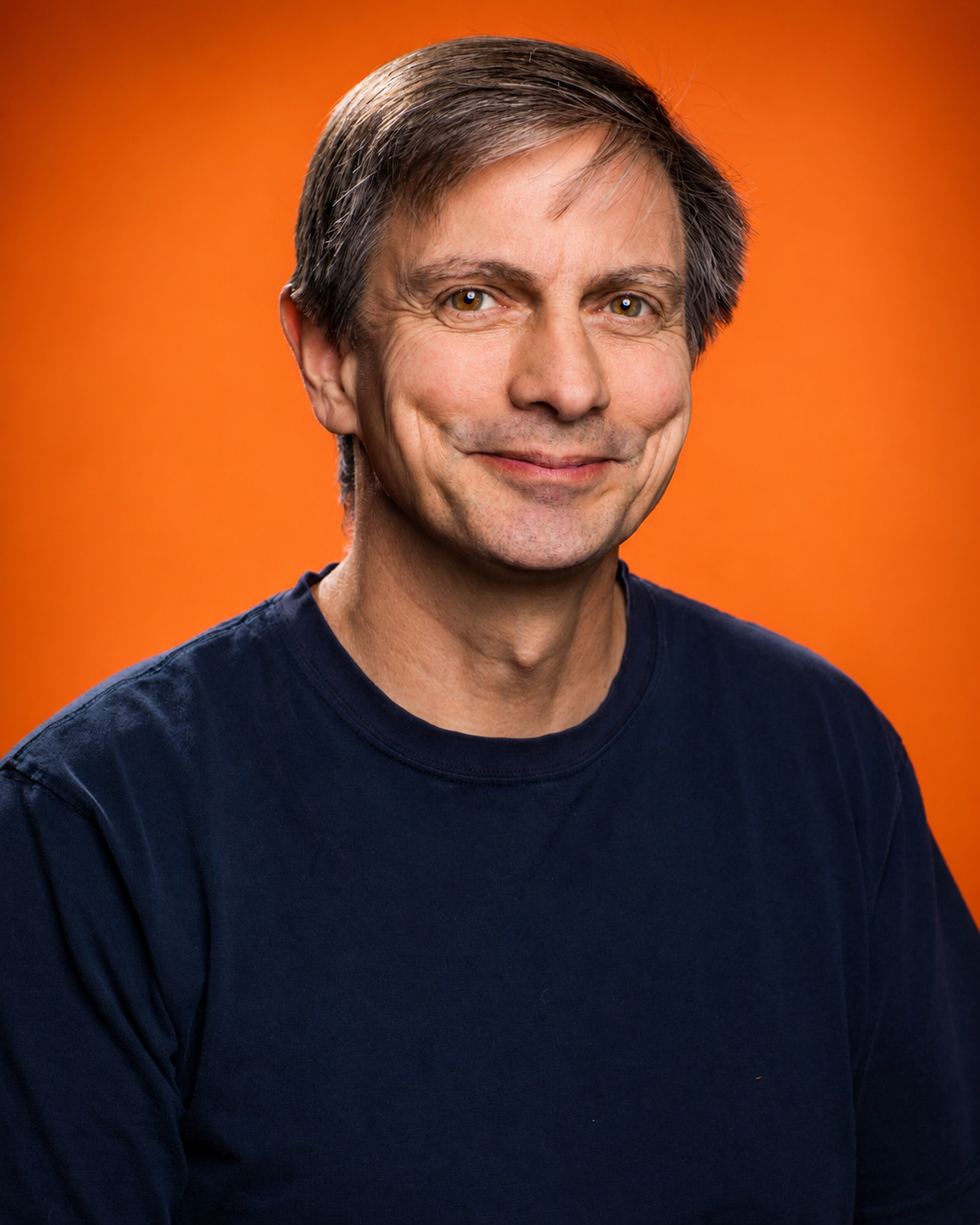 A middle-aged man with light brown hair and hazel eyes smiling, wearing a dark blue T-shirt against an orange background.