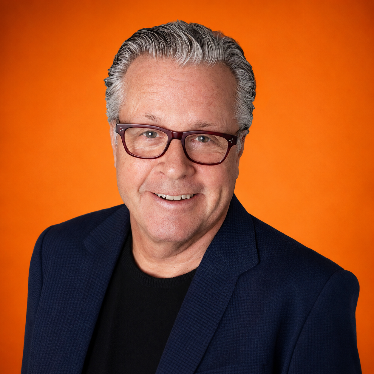 Portrait of a smiling middle-aged man with gray hair, wearing glasses, a navy blazer, and a black shirt against an orange background.