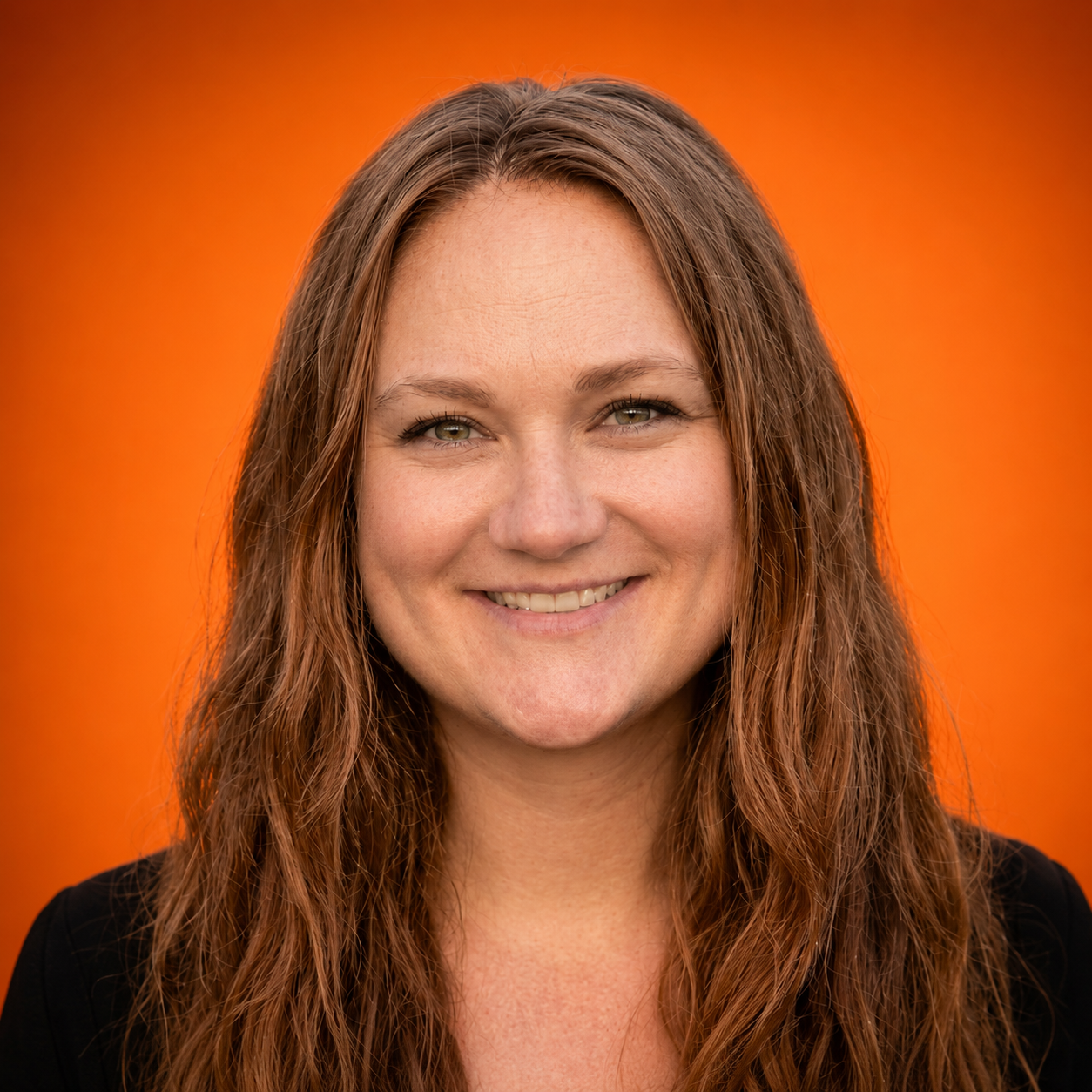A woman with long, wavy red hair and green eyes smiling against an orange background.