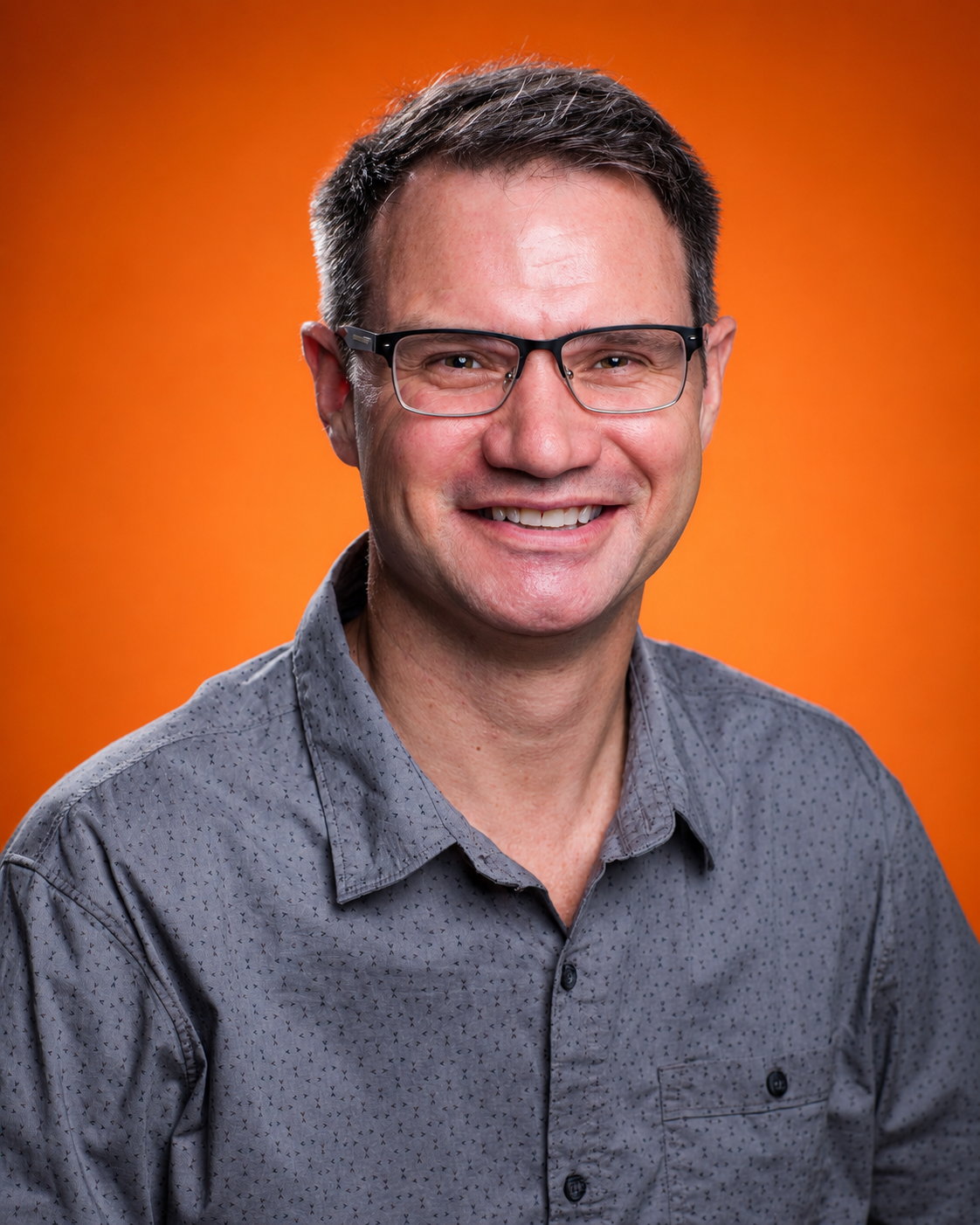 A smiling man with glasses and short dark hair, wearing a patterned collared shirt, against a plain gray background.