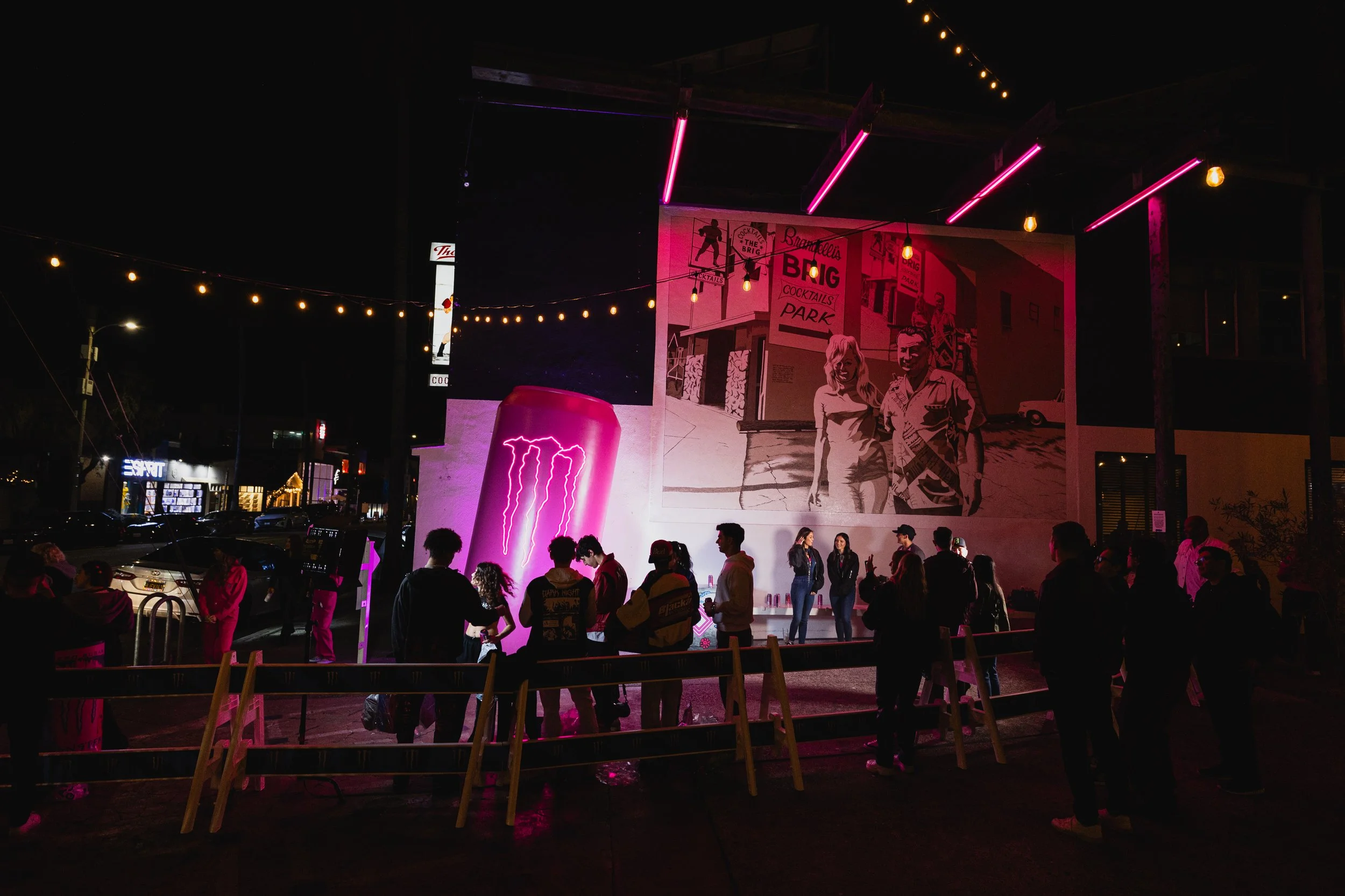 Night scene of an outdoor event with a crowd of people gathered in front of a mural and neon lights, including pink neon and string lights, with a building and parked cars in the background.