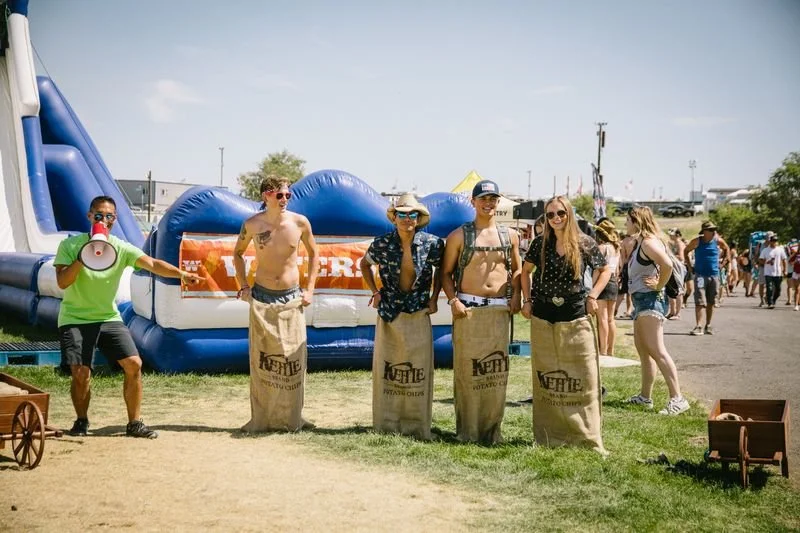 Five people standing in a line with bean bag chairs at an outdoor fair or festival, a person with a megaphone to the left, and a large inflatable slide in the background.