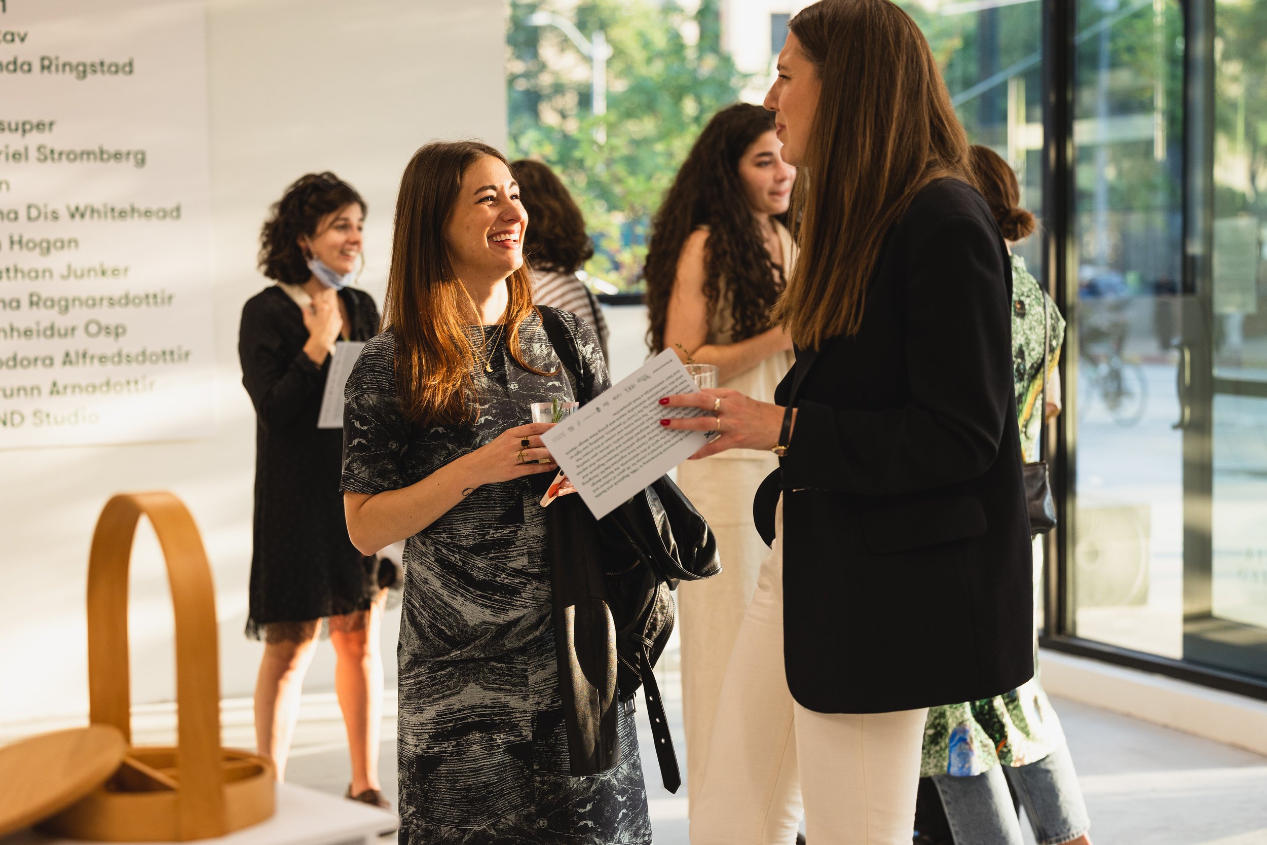 Two women smiling and talking inside a modern building with large glass windows, with other women in the background engaged in conversation.