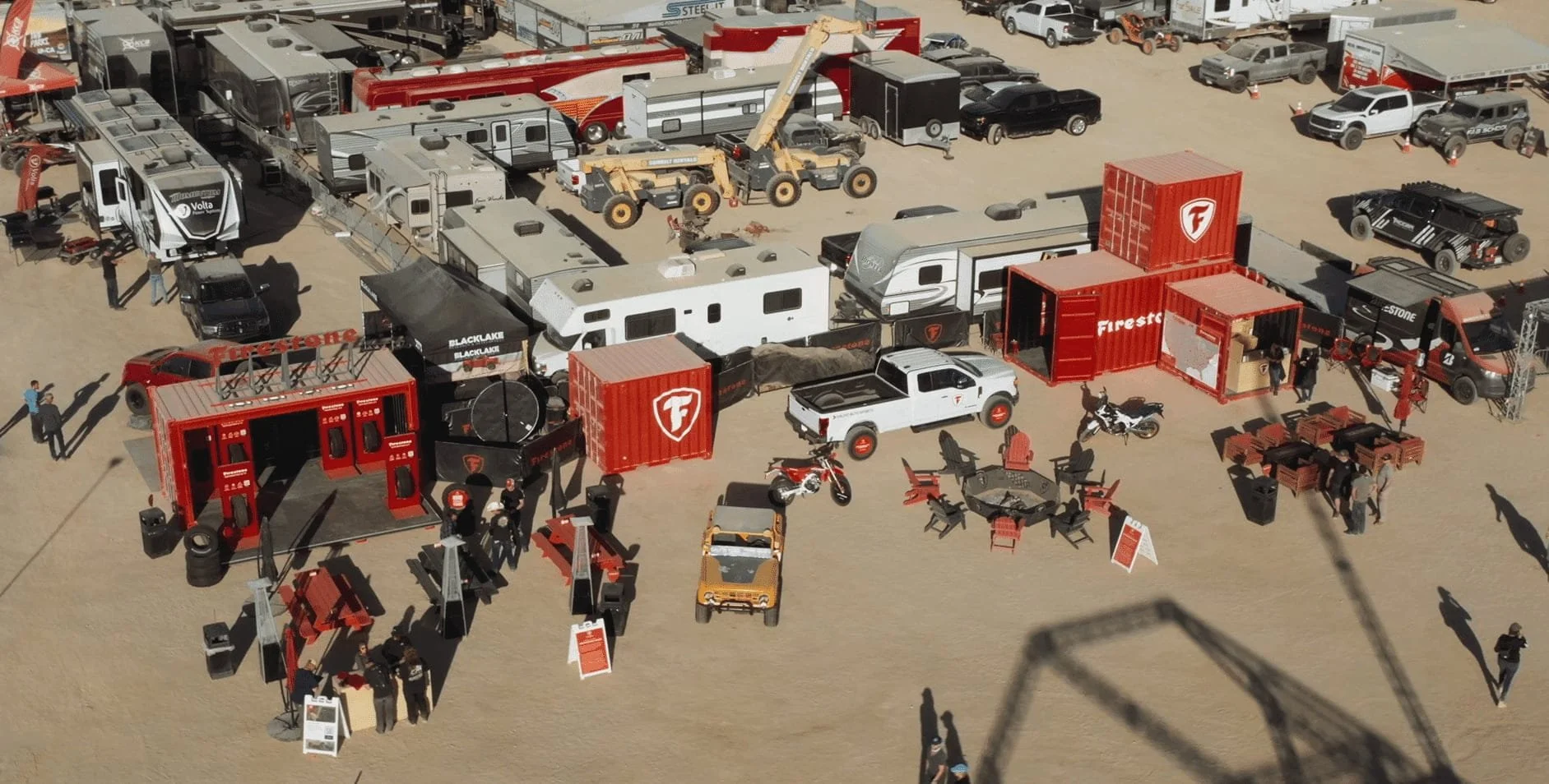 High-angle aerial view of a Firestone brand event setup featuring red shipping containers, vehicles, trailers, and seating areas in a desert lot.