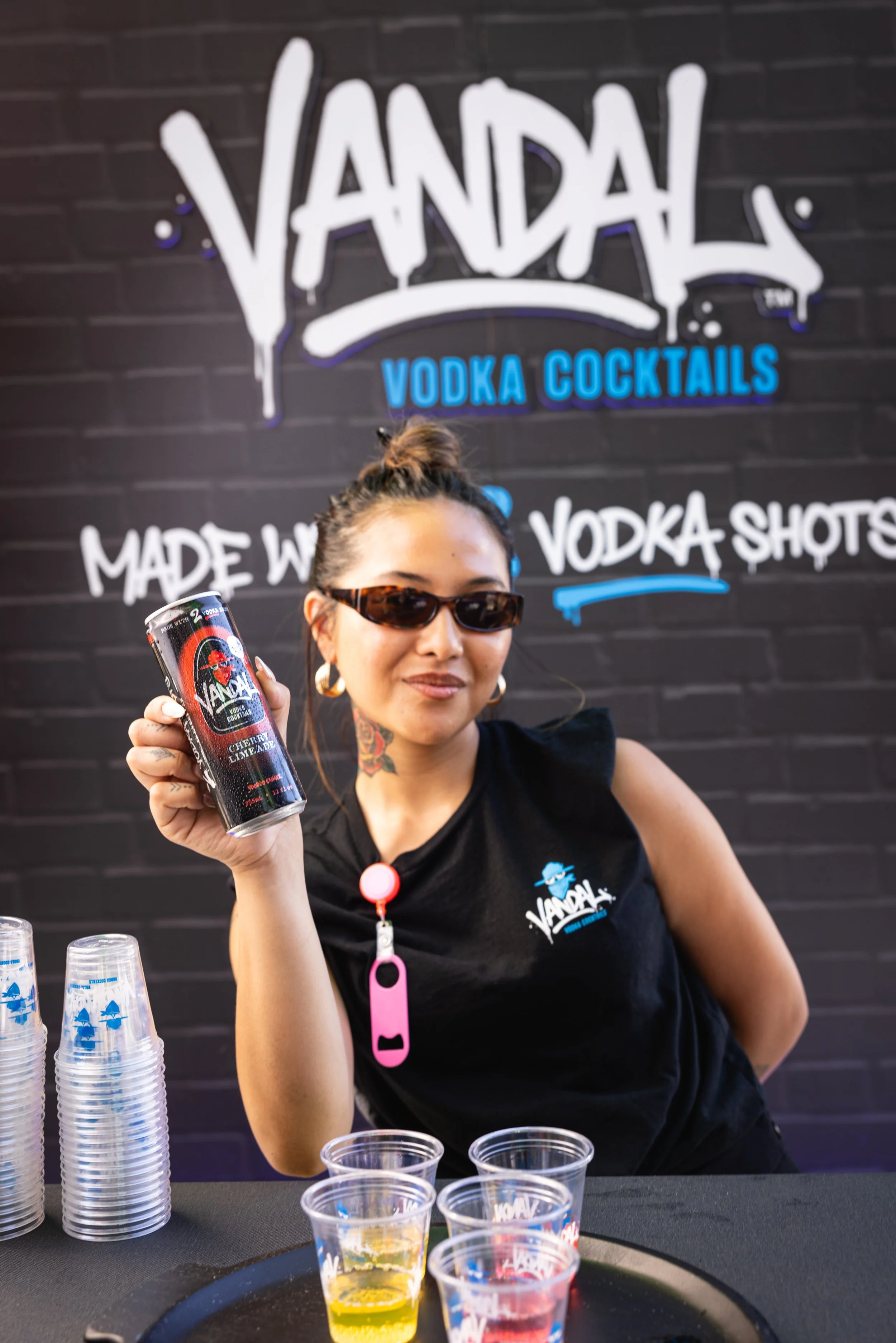 A woman with sunglasses holding a can of Vandal vodka cocktail at a vendor booth. Behind her is a sign that reads "Vandal Vodka Cocktails" on a black brick wall. The booth has cups and containers for serving drinks.
