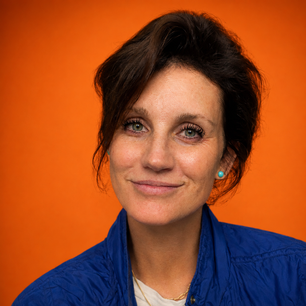 Close-up portrait of a smiling woman with short dark hair, green eyes, wearing blue earrings and a blue jacket, with an orange background.
