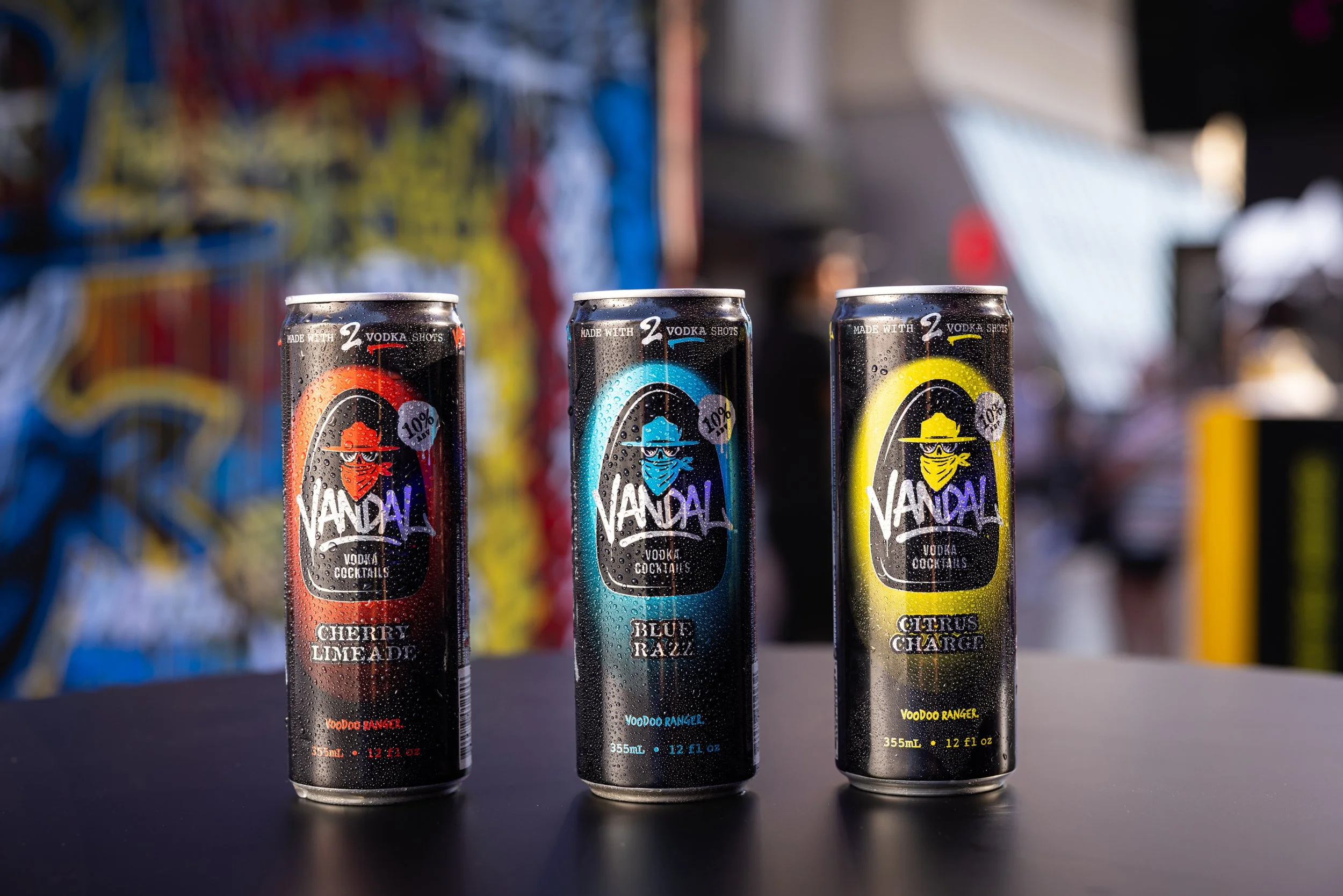 Three energy drink cans labeled 'Vandal' with different flavors: cherry limeade, blue razz, and citrus charge, placed on a dark surface with a blurry colorful background.