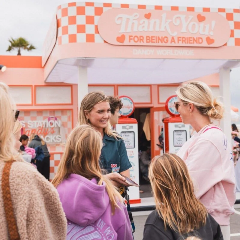 A group of women and children standing outside a pink and orange ice cream stand with a sign that says "Thank You! For Being a Friend" at an amusement park or fair.