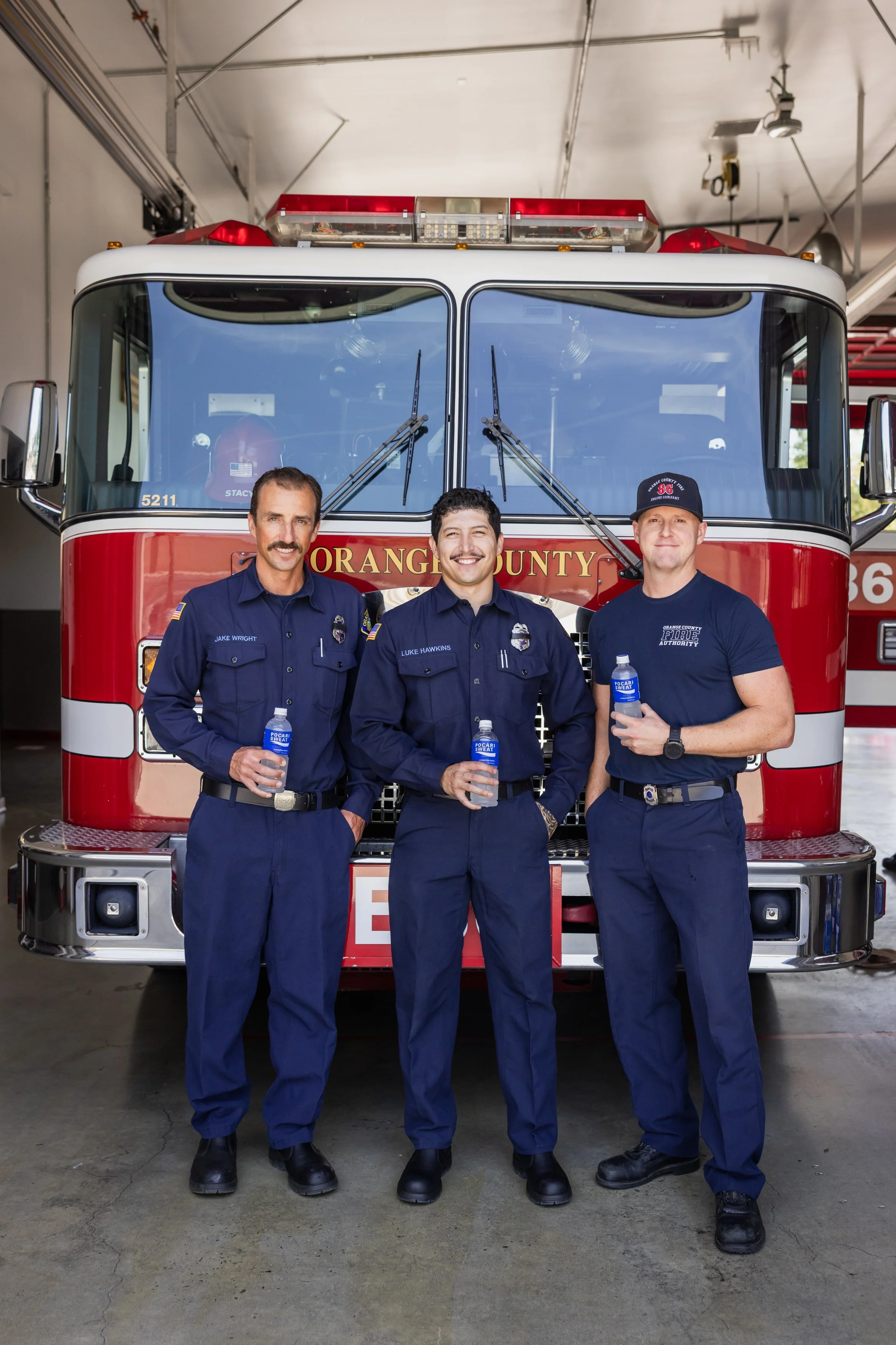 Three firefighters wearing navy uniforms and holding water bottles, standing in front of a red and white fire truck with 
