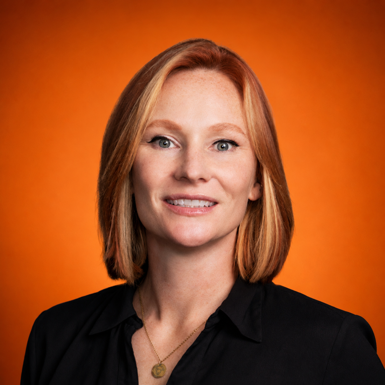 A woman with shoulder-length red hair, fair skin, and blue eyes smiling against a warm orange background.