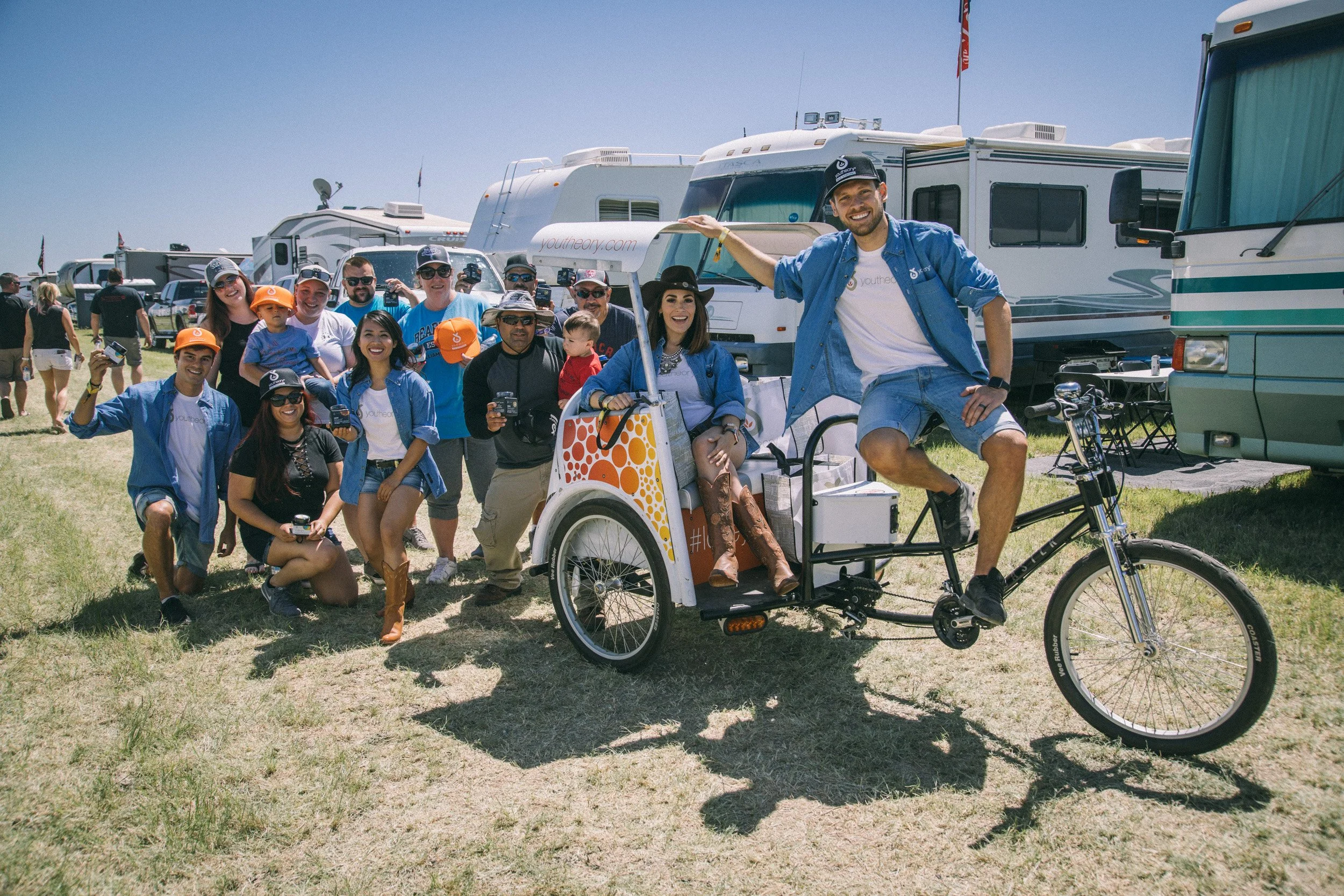 A group of people gathered at a campground with RVs and trailers in the background, including a man riding a bicycle with a pedal-assisted tricycle and a woman sitting on it, all smiling.