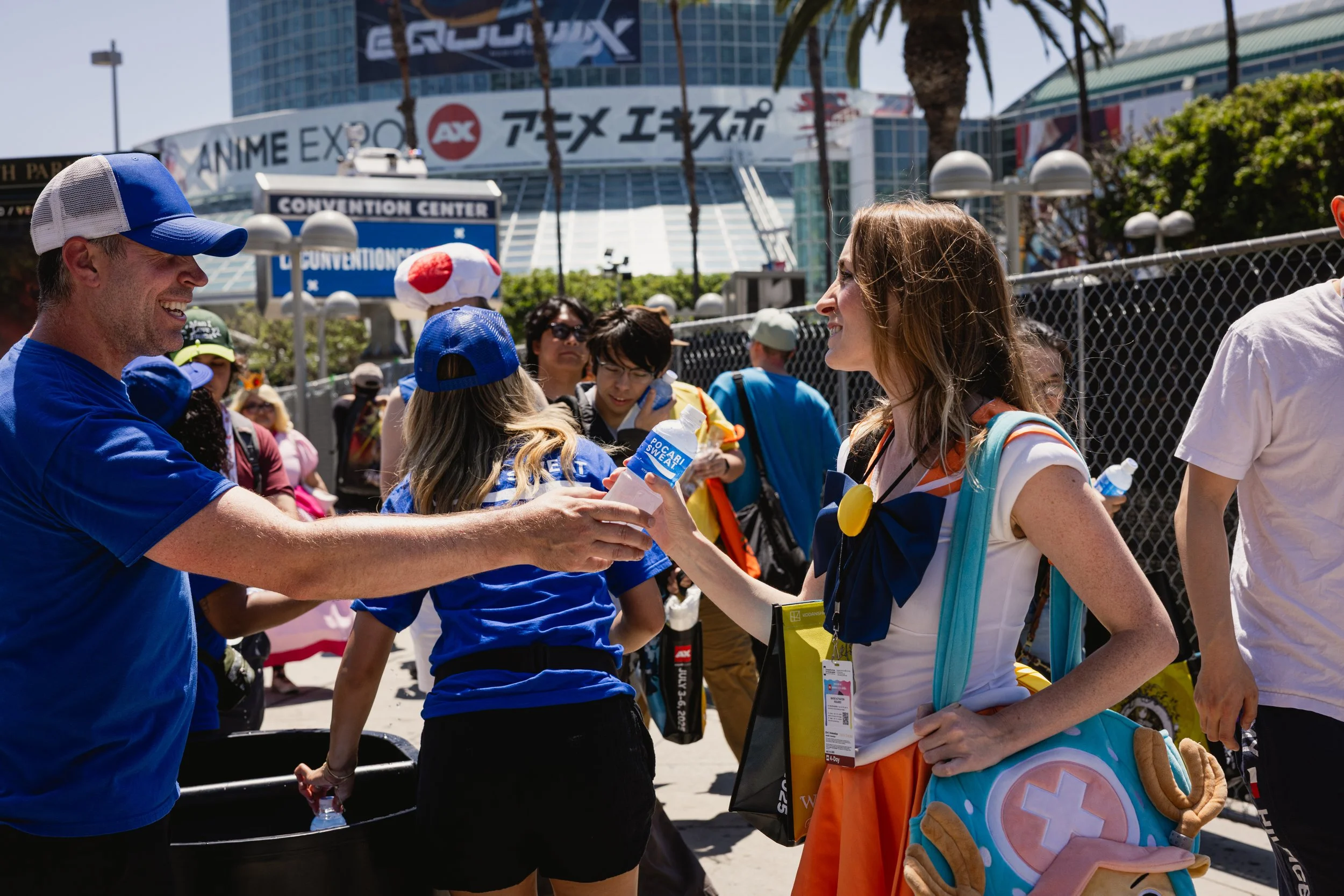 A group of people at Anime Expo in front of the Los Angeles Convention Center. One woman dressed as a cartoon character is shaking hands with a man in a blue shirt and hat, while other attendees mingle nearby.