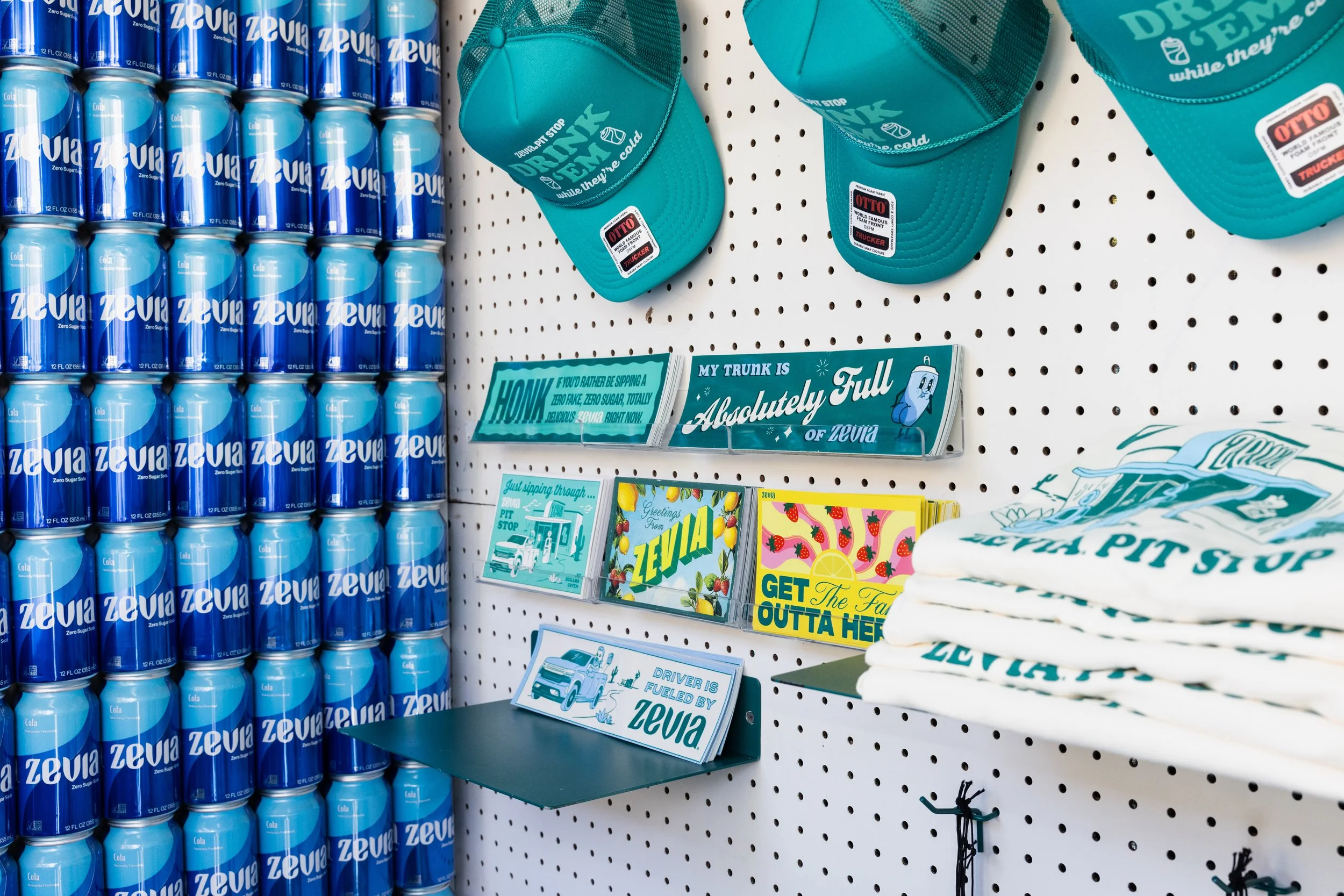 A display of Zevia zero sugar soda cans, teal hats with 'Drink Zevia while they’re cold' written on them, and colorful pamphlets about Zevia on a white pegboard.