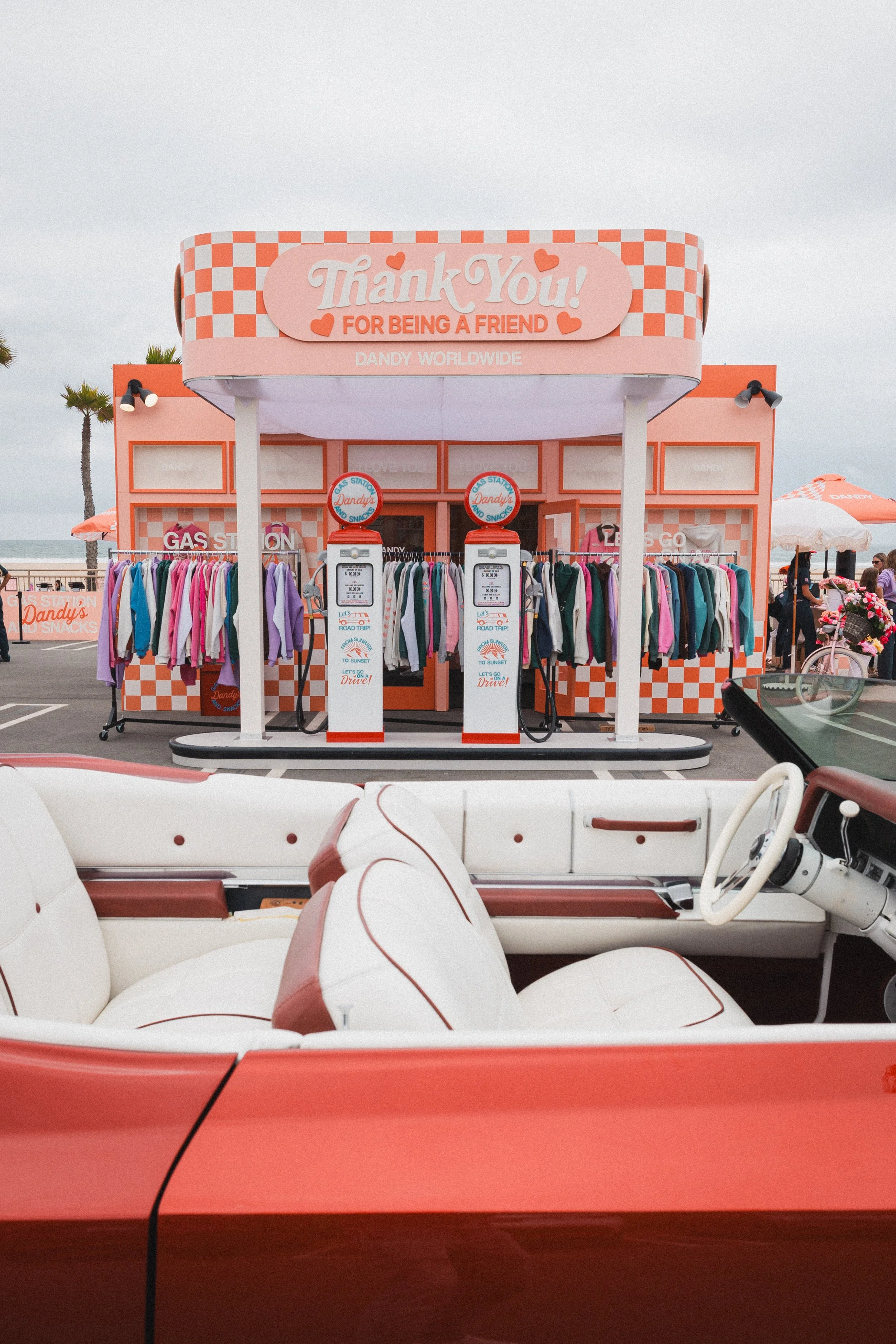A pink and orange checkered snack bar with a sign reading 'Thank You! For Being A Friend' at a beachside location. There are vintage gas pumps and a rack of colorful jackets in front, with the ocean and palm trees in the background.
