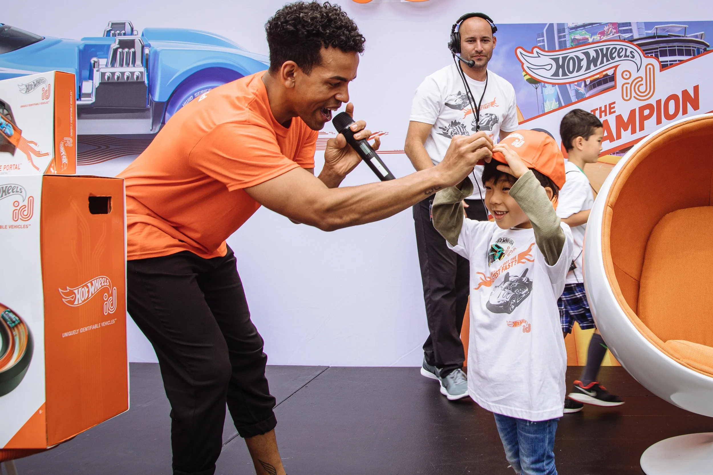 A man in an orange shirt is placing a cap on a young boy's head at a Hot Wheels event. There is another man with a headset and microphone in the background, along with several children and promotional displays for Hot Wheels.