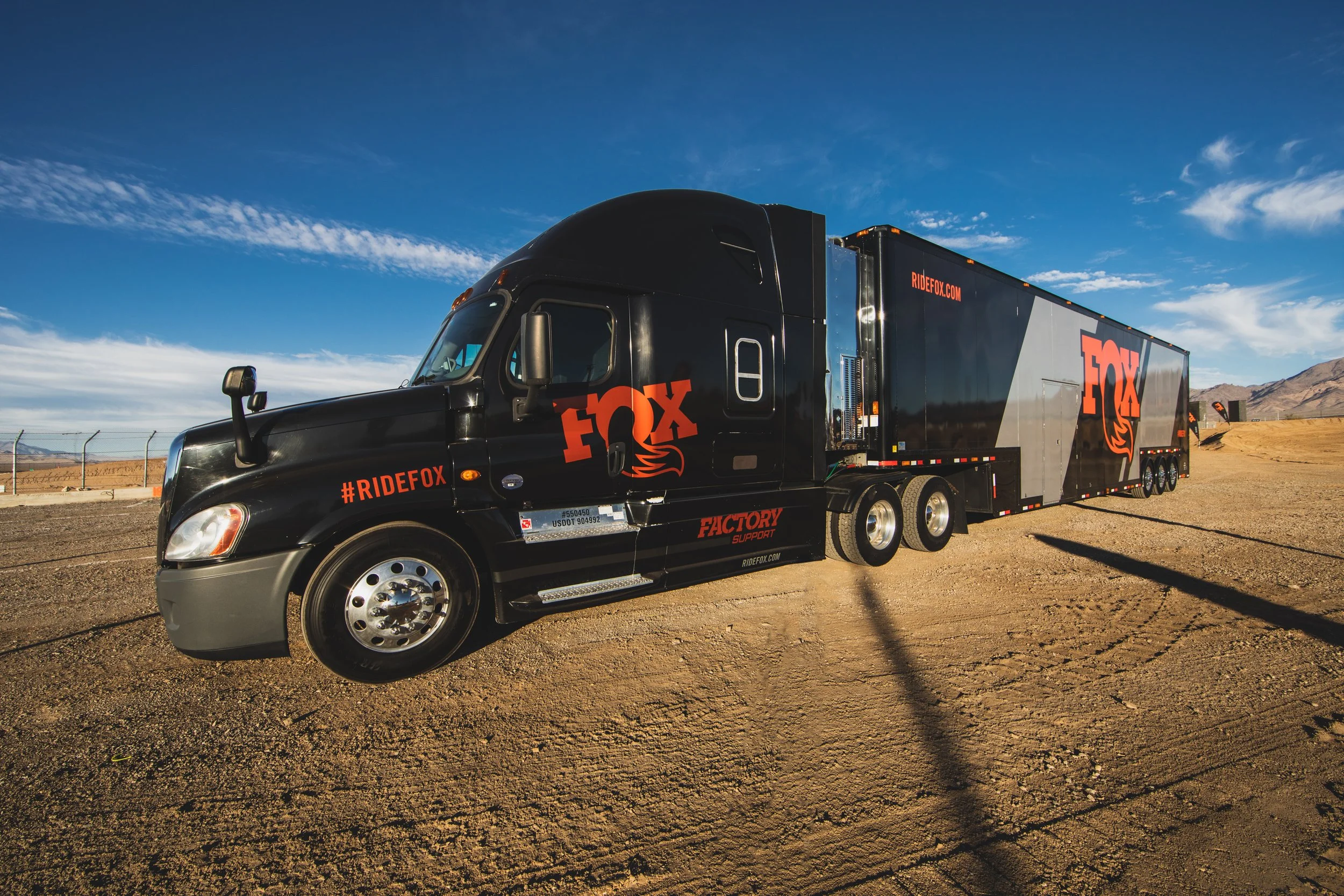 A black racing truck with red and orange branding, parked on dirt terrain with mountains and a blue sky in the background.