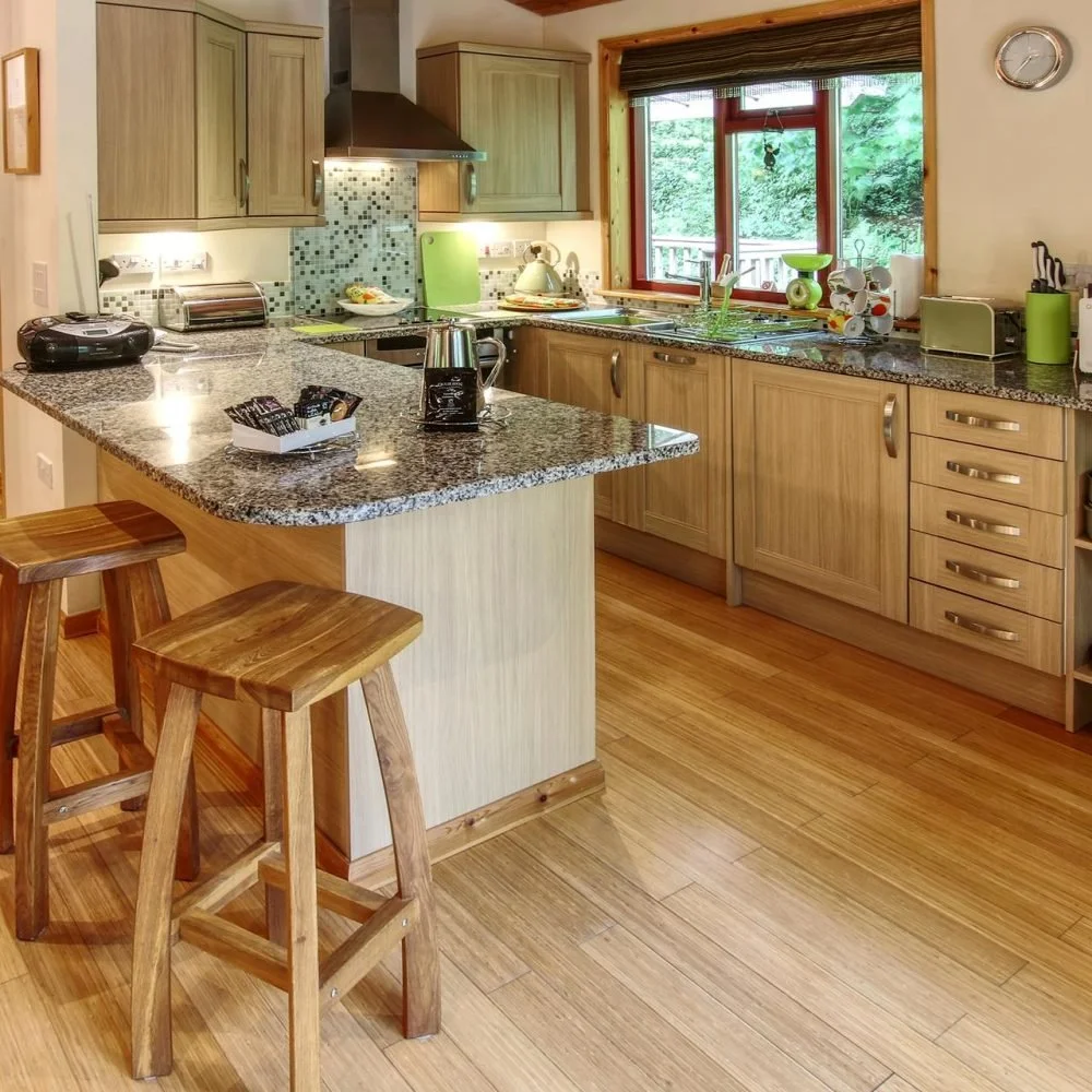A cozy kitchen with wooden cabinets, granite countertops, and a large window overlooking a green outdoor space. The counter has kitchen appliances and utensils, and there are two wooden stools at a breakfast bar.