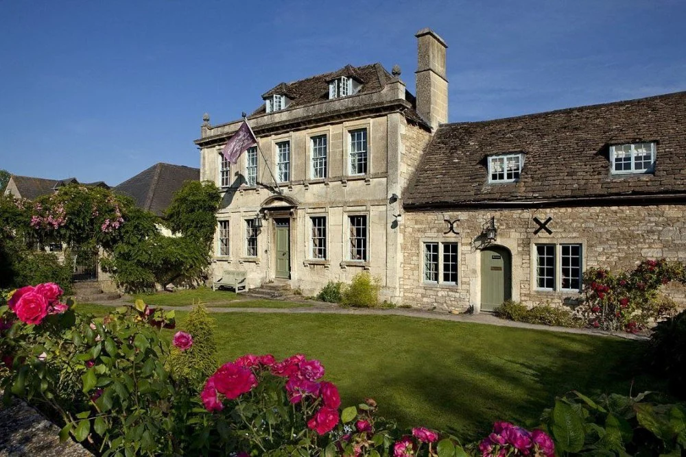 A historic stone house with multiple windows and a rooftop, surrounded by a well-maintained lawn and colorful flowers, under a clear blue sky.