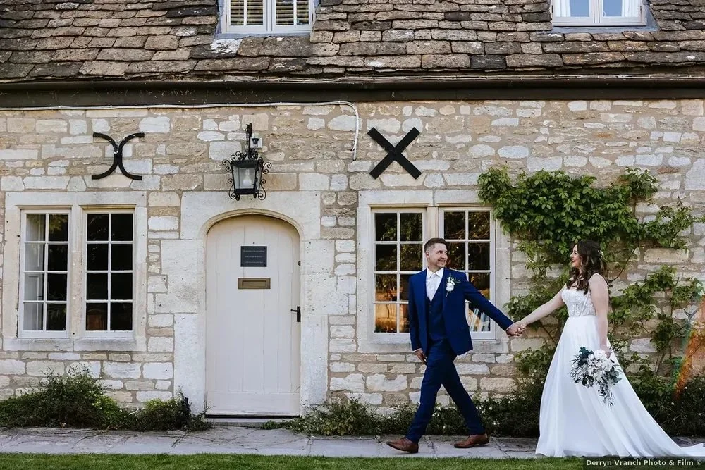 A bride and groom holding hands and walking in front of a stone house with large windows and decorative symbols on the wall.