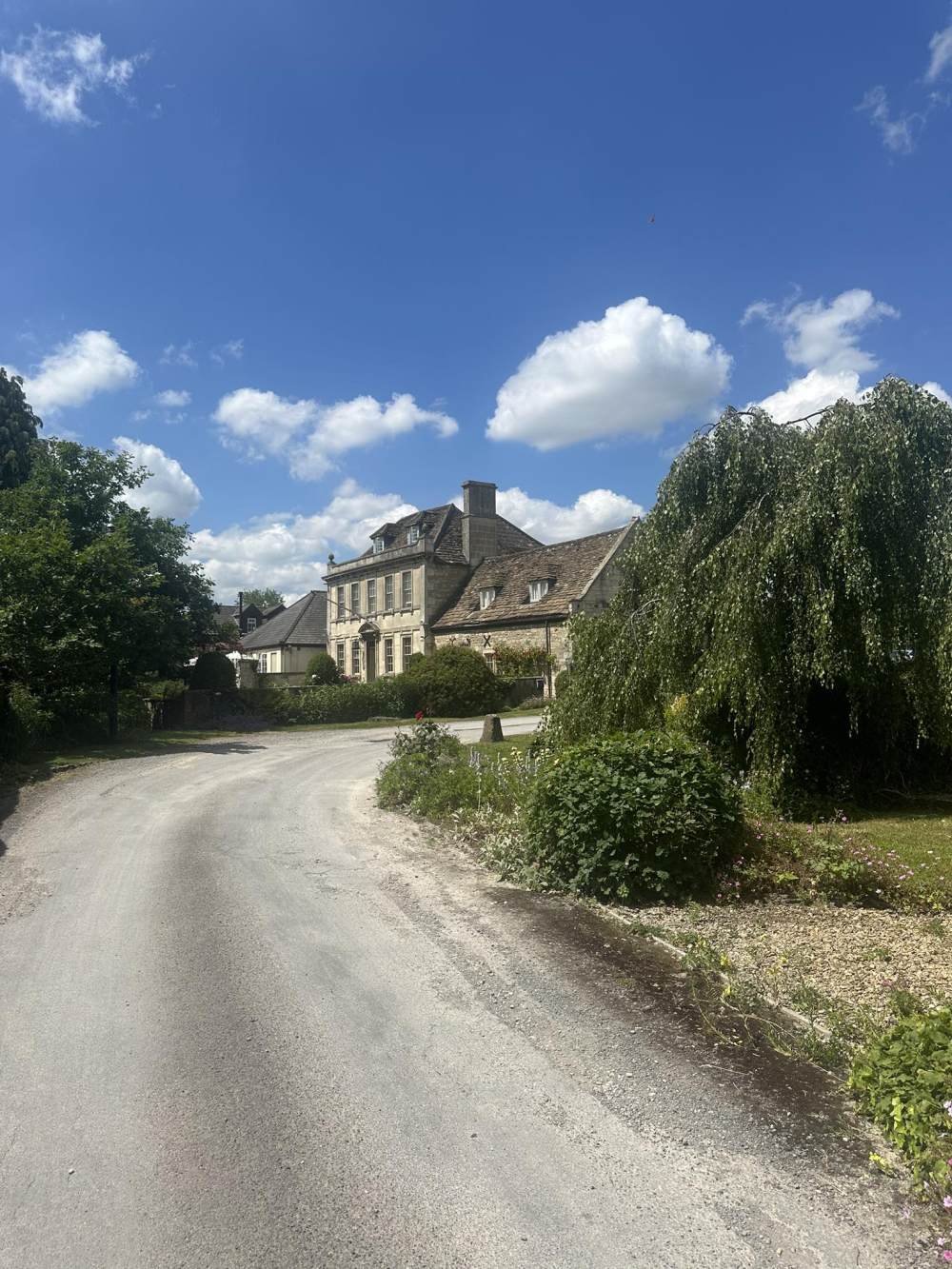 A gravel road curves past a large, historic stone house with multiple stories, set against a backdrop of a bright blue sky with scattered white clouds. There are trees and bushes along the roadside.