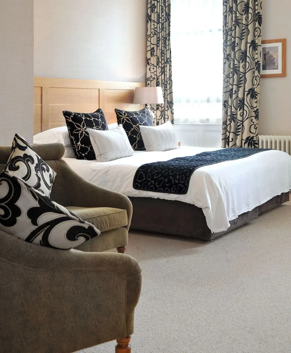 A neatly made bed with black and white patterned pillows and a black throw blanket, next to a beige armchair with matching patterned pillows, in a well-lit bedroom with curtains and a framed picture on the wall.