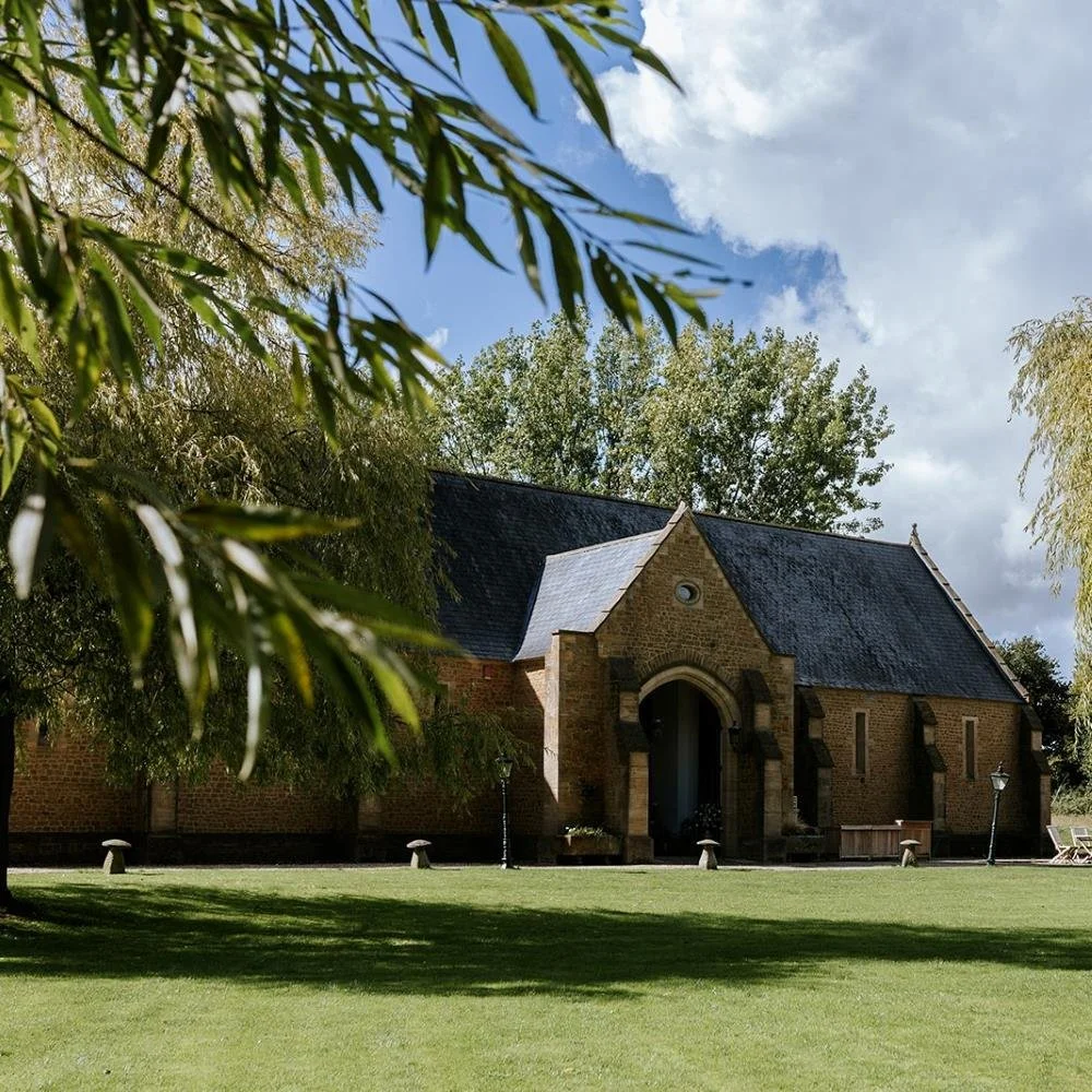 A brick church building with a pointed roof, surrounded by a grassy lawn and trees, under a partly cloudy sky.