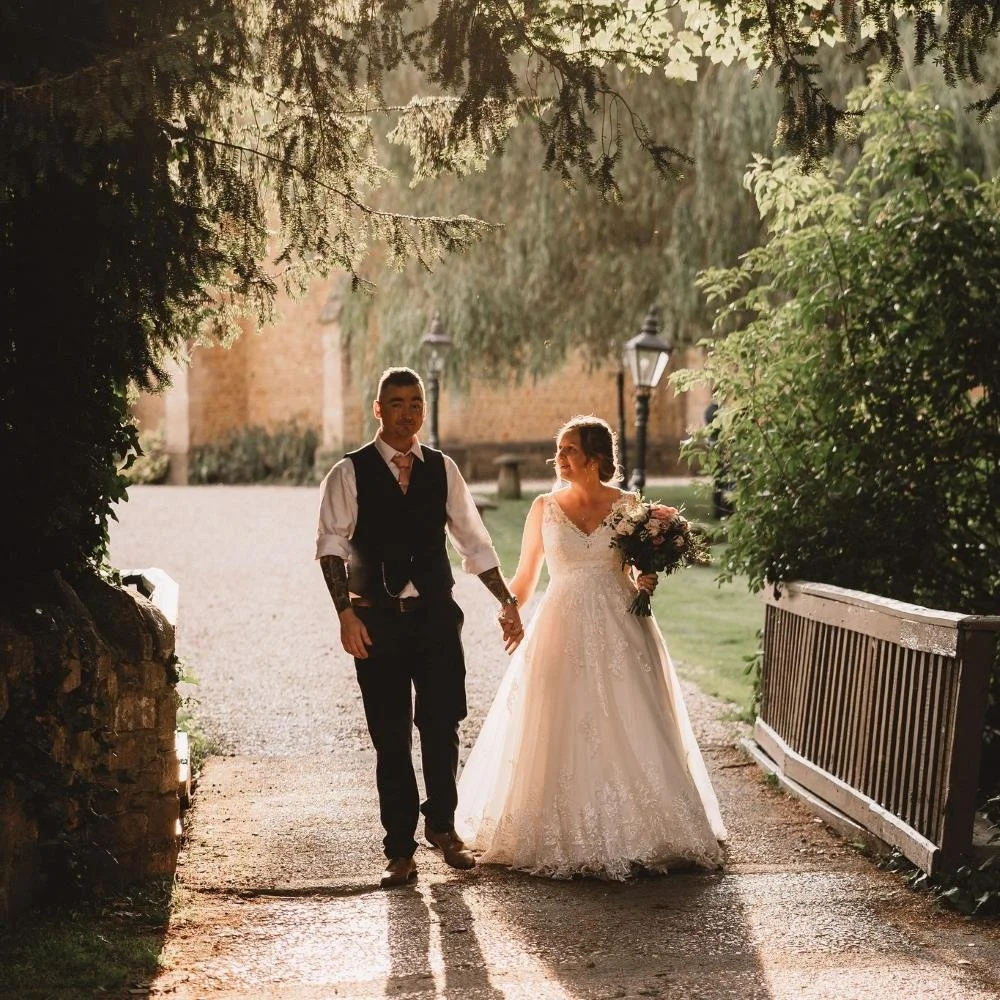 A bride and groom walking hand in hand outside on a sunny day, with trees and lanterns in the background. The bride is holding a bouquet of flowers.